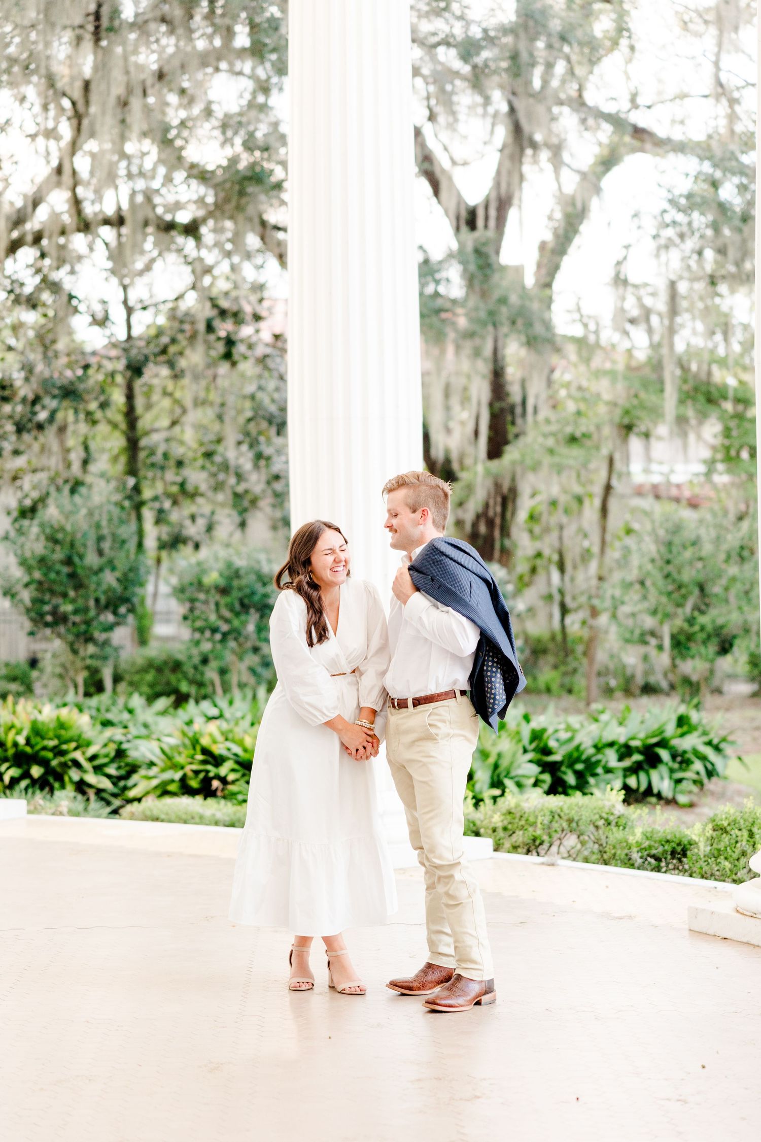 A couple holds hands and smiles near a white column in a lush garden, woman in white dress, man in blazer.