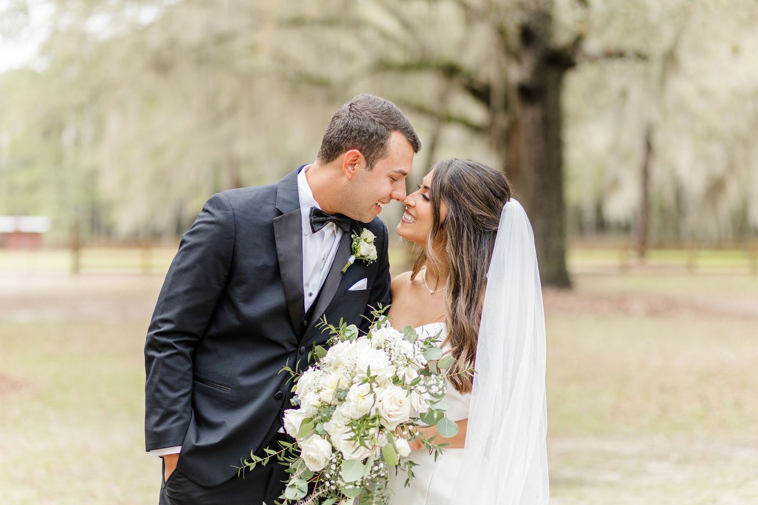 Elegant wedding couple shares intimate moment while holding white floral bouquet outdoors.