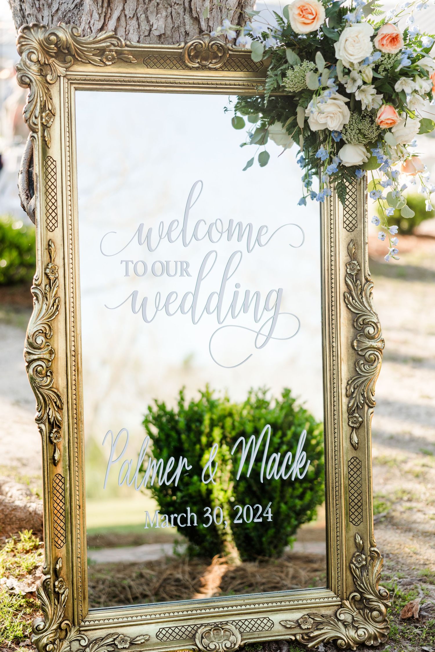An ornate gold mirror wedding welcome sign decorated with fresh flowers.