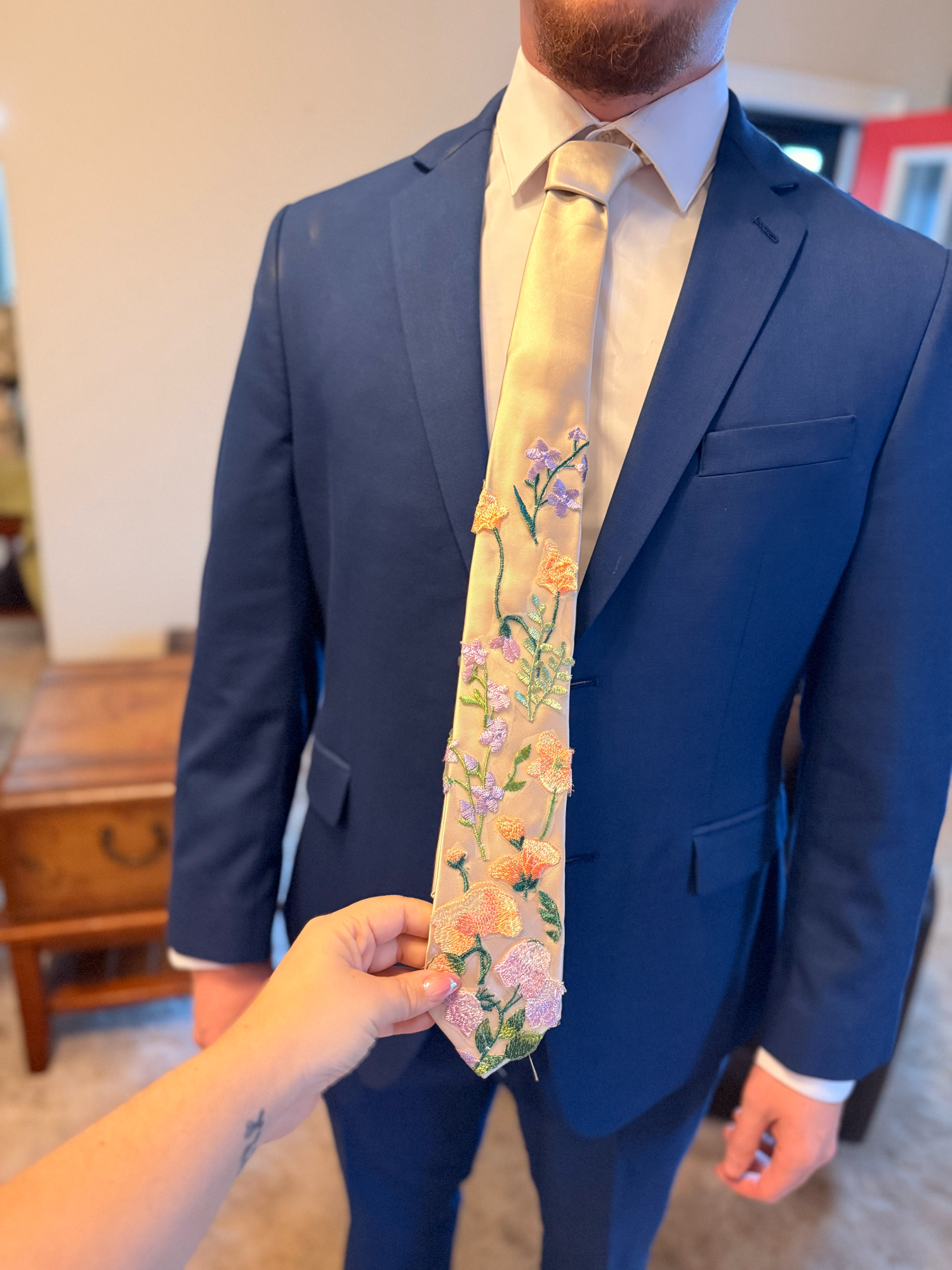 Man in navy blue suit wearing a floral embroidered tie with pink, yellow, and purple flowers on cream fabric.
