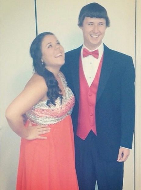 Couple dressed in formal prom attire, woman in coral gown and man in black tuxedo with red vest and bow tie.