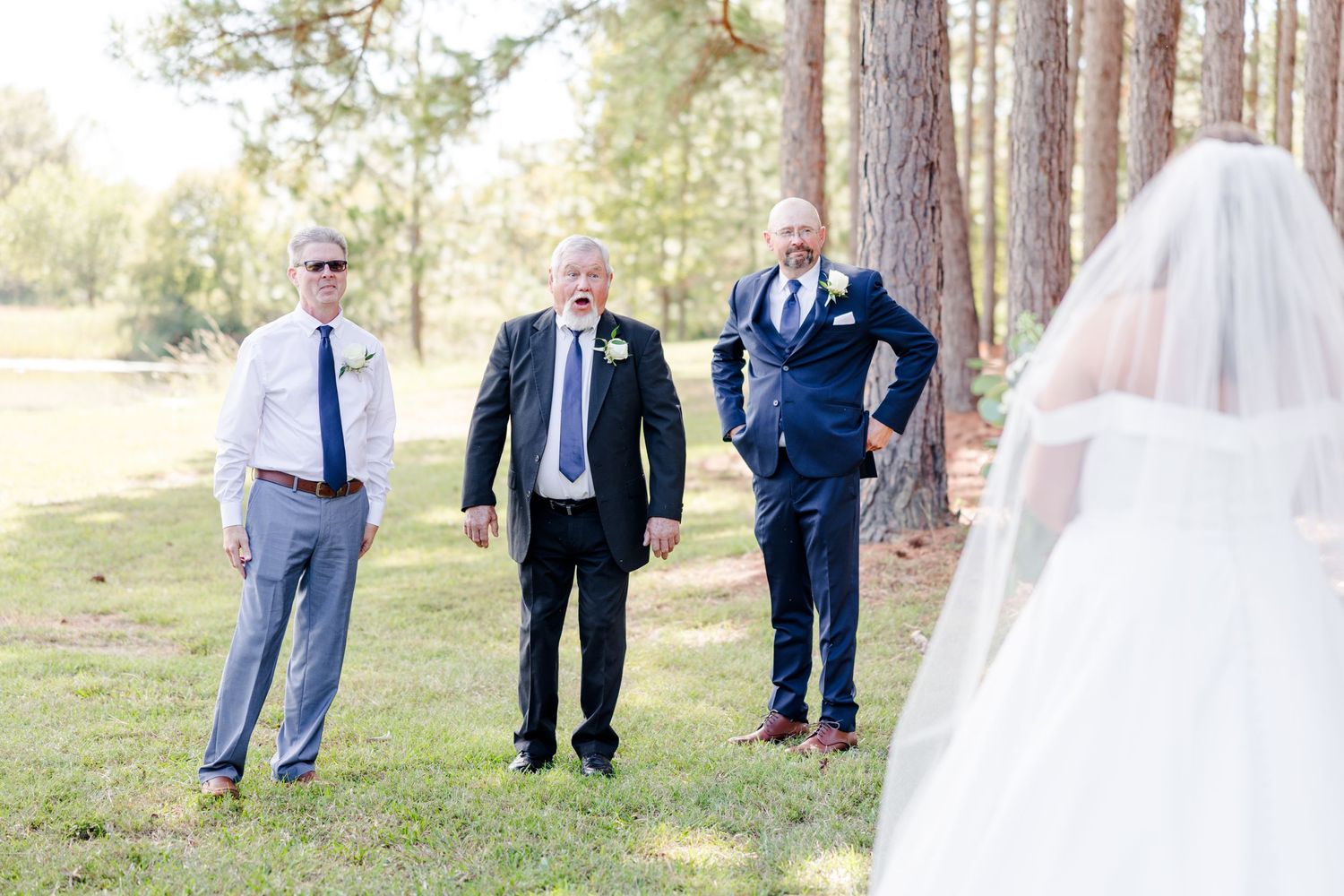 Wedding party members stand spaced apart in a line outdoors among tall pine trees.