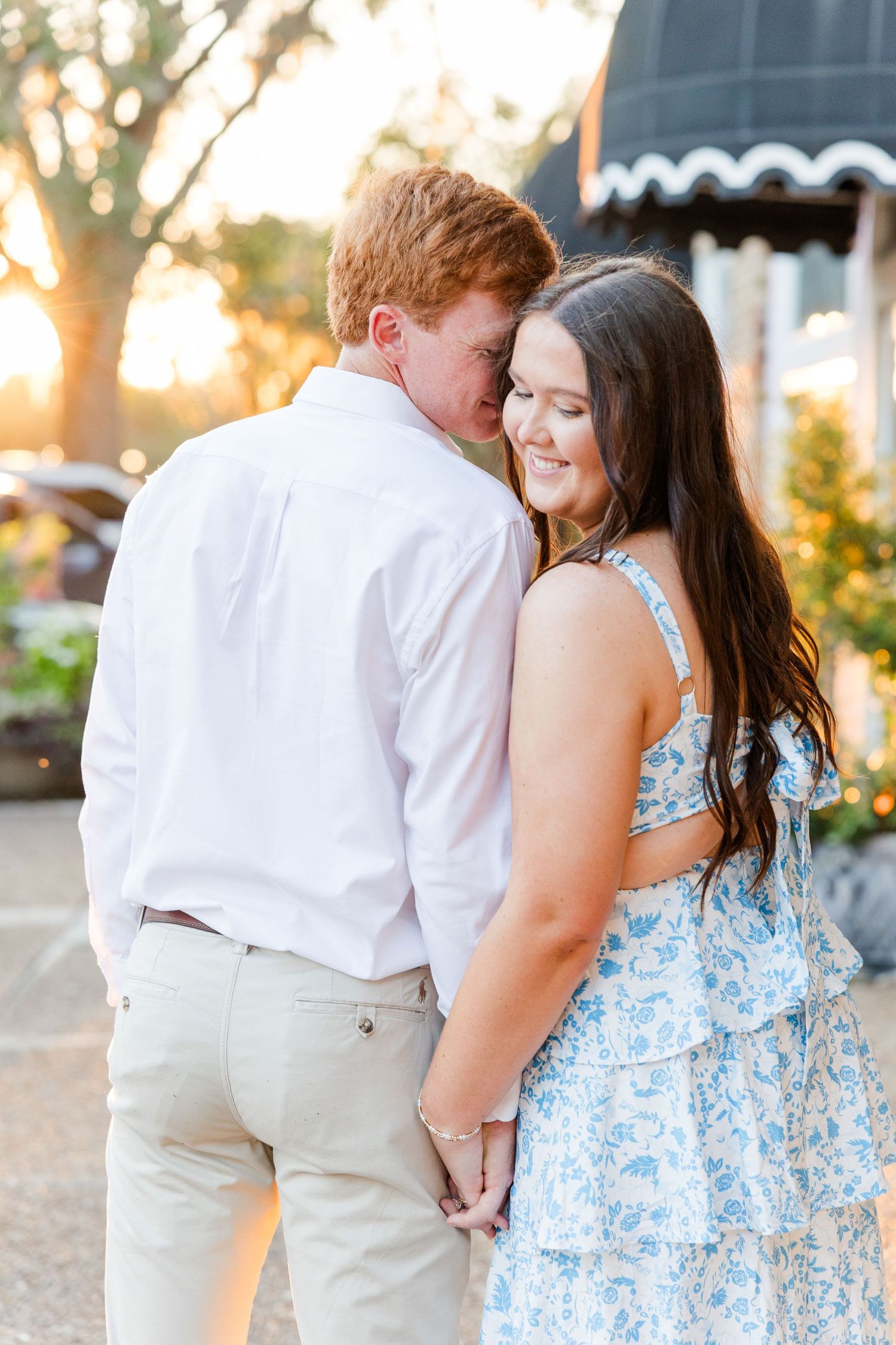 Couple holding hands at golden hour, woman in blue floral dress smiling as man whispers in her ear outdoors.