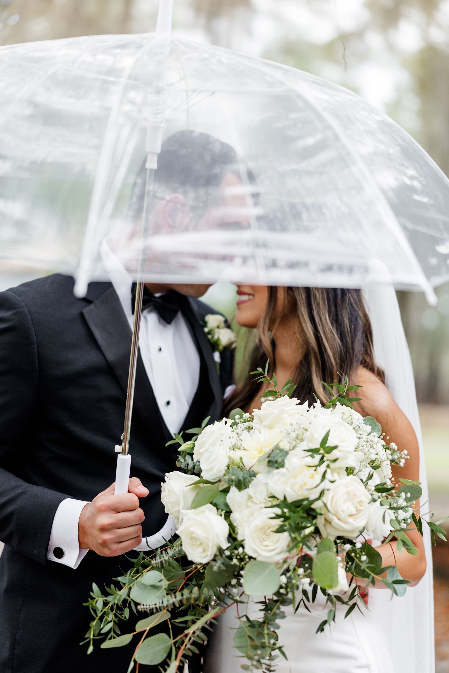 A romantic rainy day wedding photo with the couple sharing a clear umbrella and holding white roses.