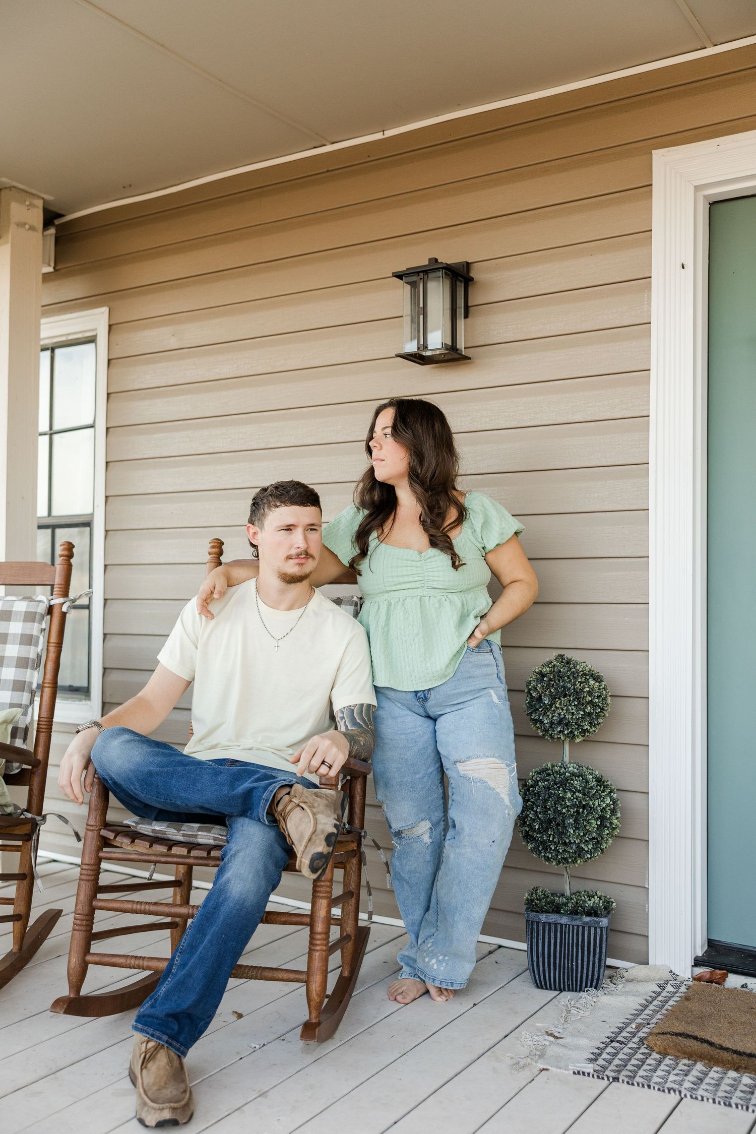 Couple relaxing on a porch, man sitting in rocking chair, woman in green top leaning against house siding.