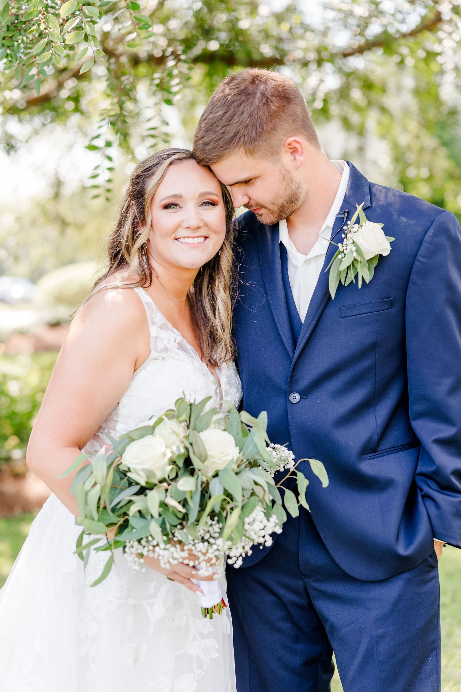 Romantic outdoor wedding couple in navy suit and white dress share intimate moment under green trees.