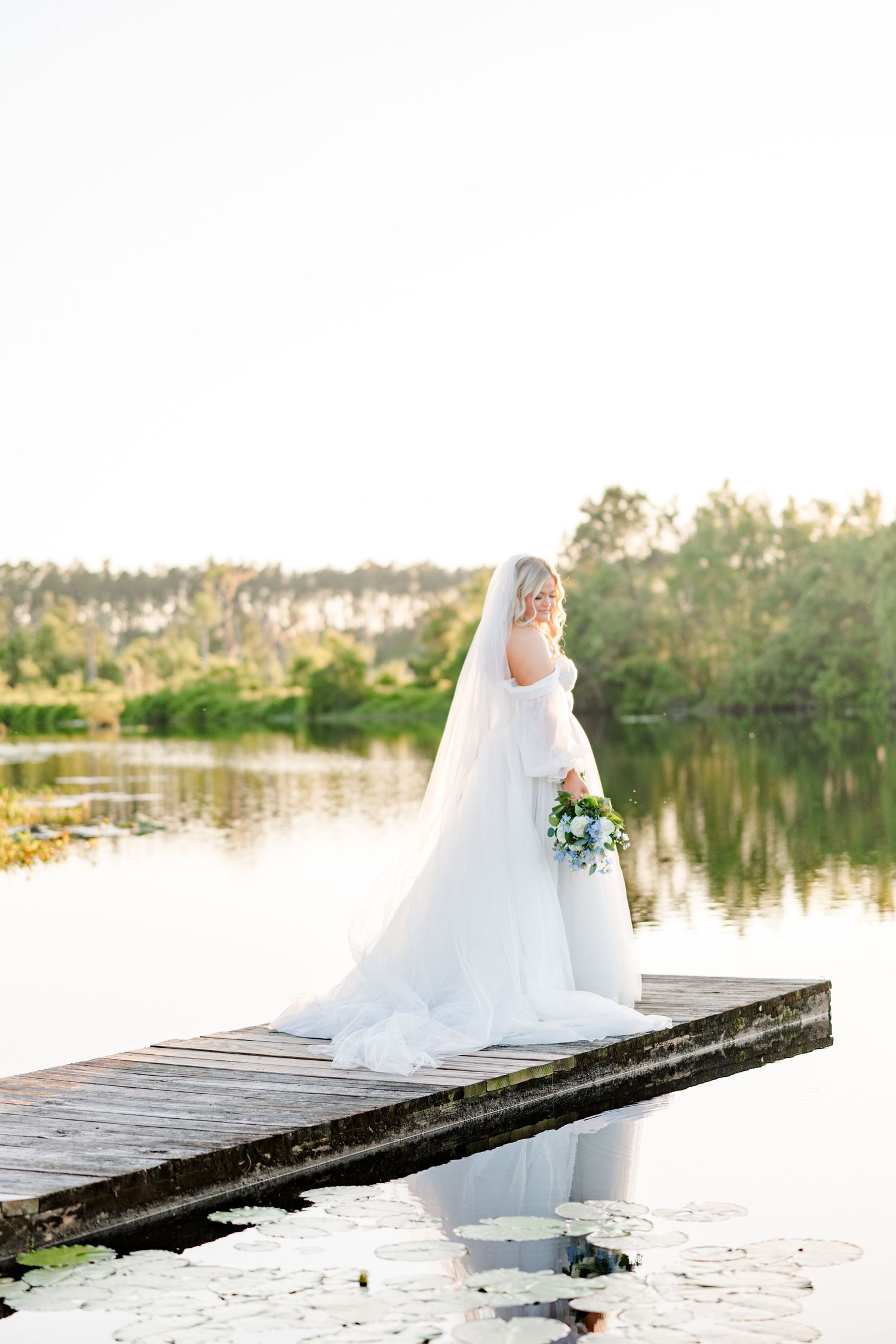 A bride in a flowing white dress stands on a wooden dock by a tranquil lake at sunset holding a bouquet.