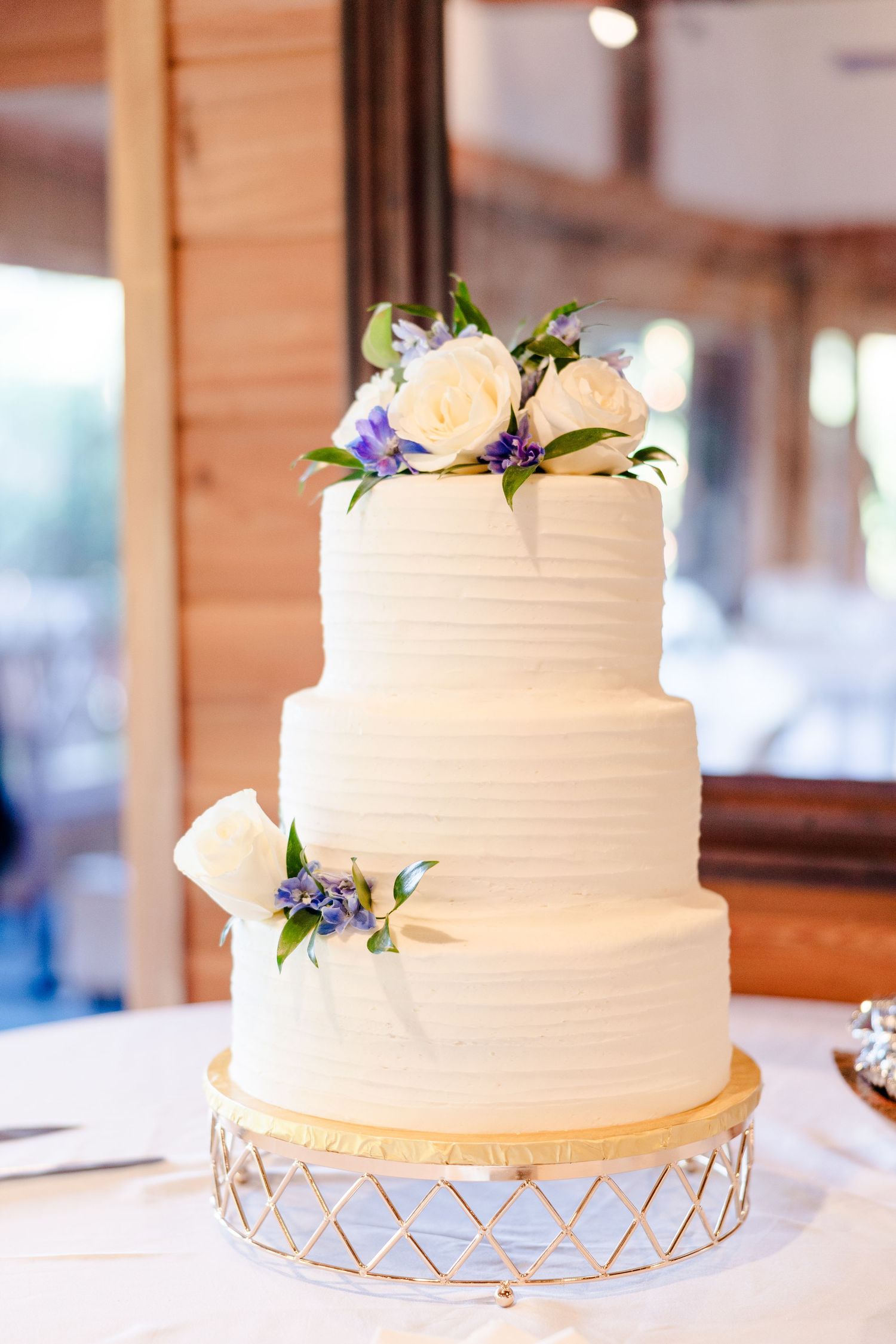 A three-tiered white wedding cake adorned with purple flowers sits on a gold stand.
