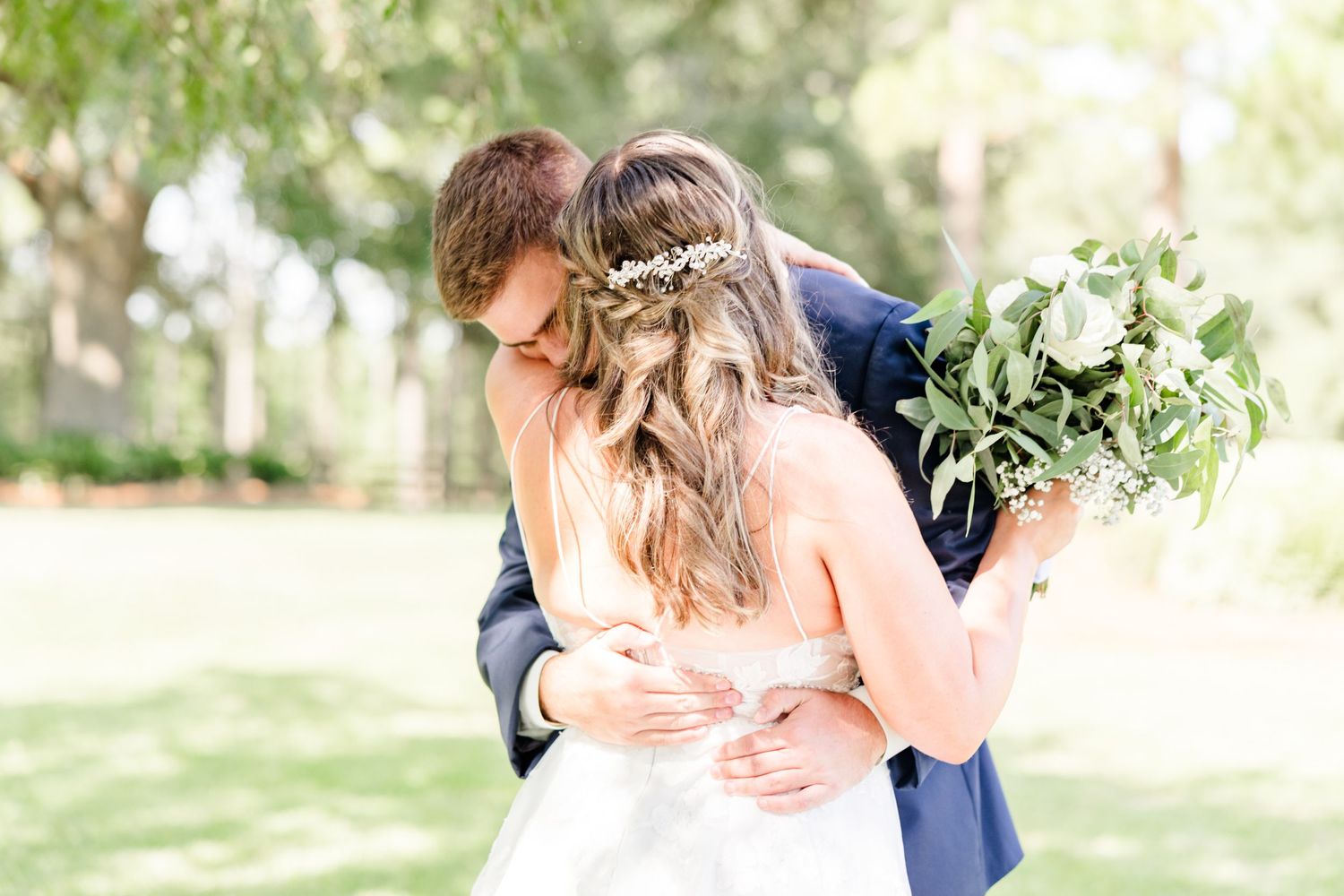 Wedding couple shares tender moment under trees with bride's lace dress flowing.
