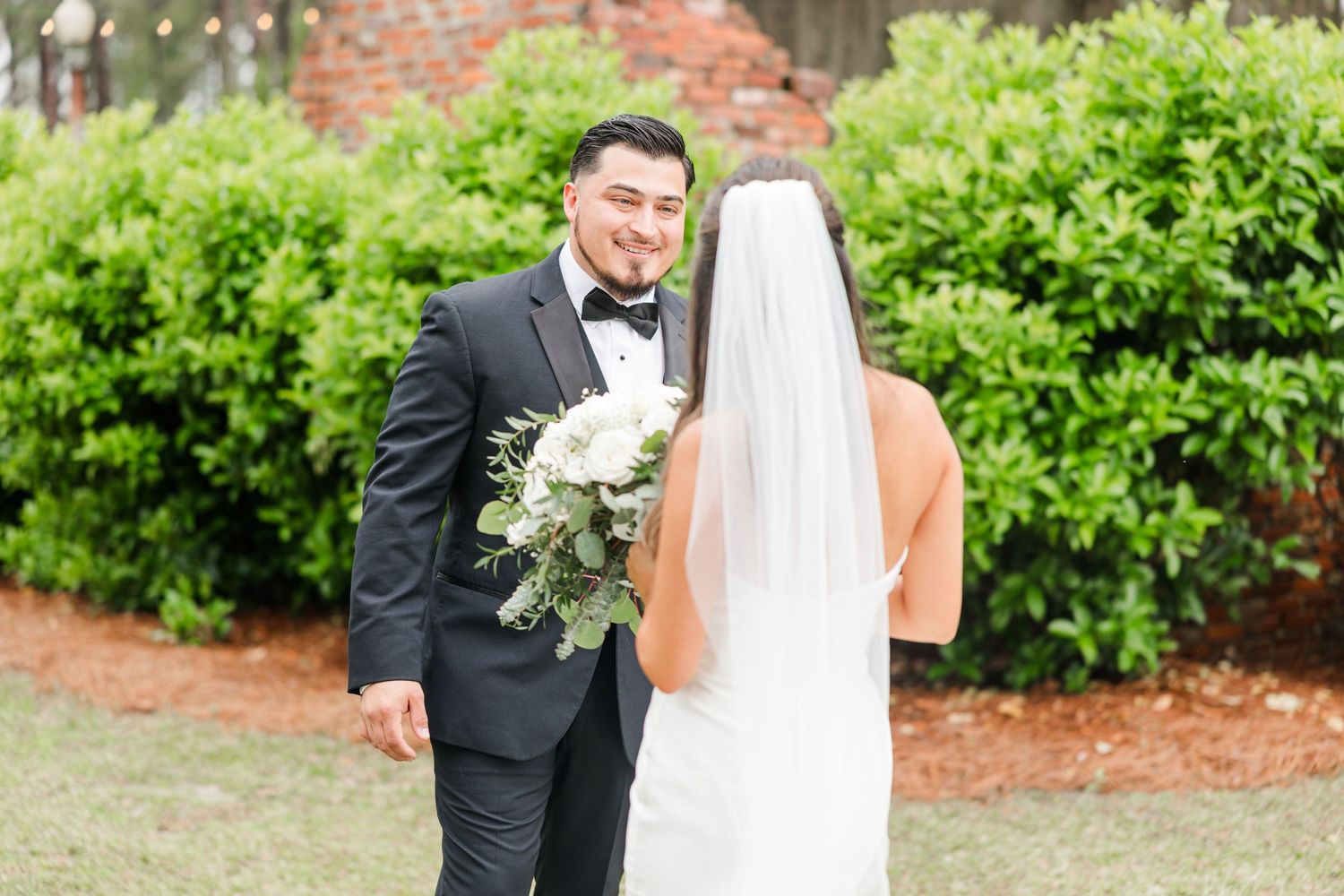 Elegant wedding couple poses in front of lush green bushes and brick wall.