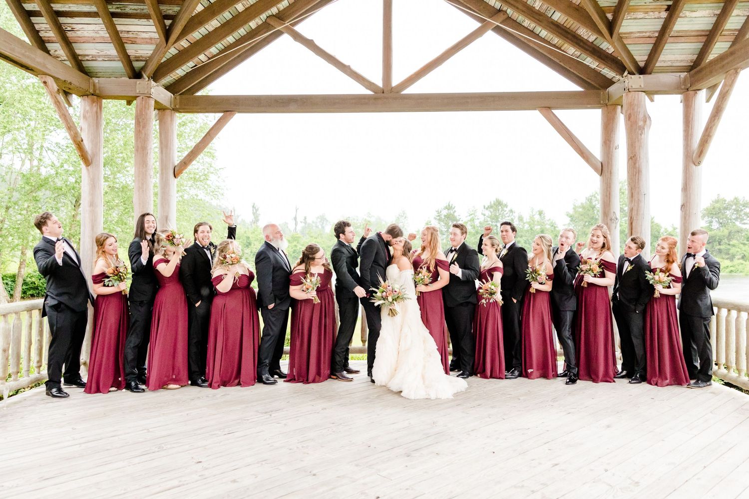 Wedding party in burgundy dresses and black suits under a wooden pavilion, celebrating with the bride and groom.