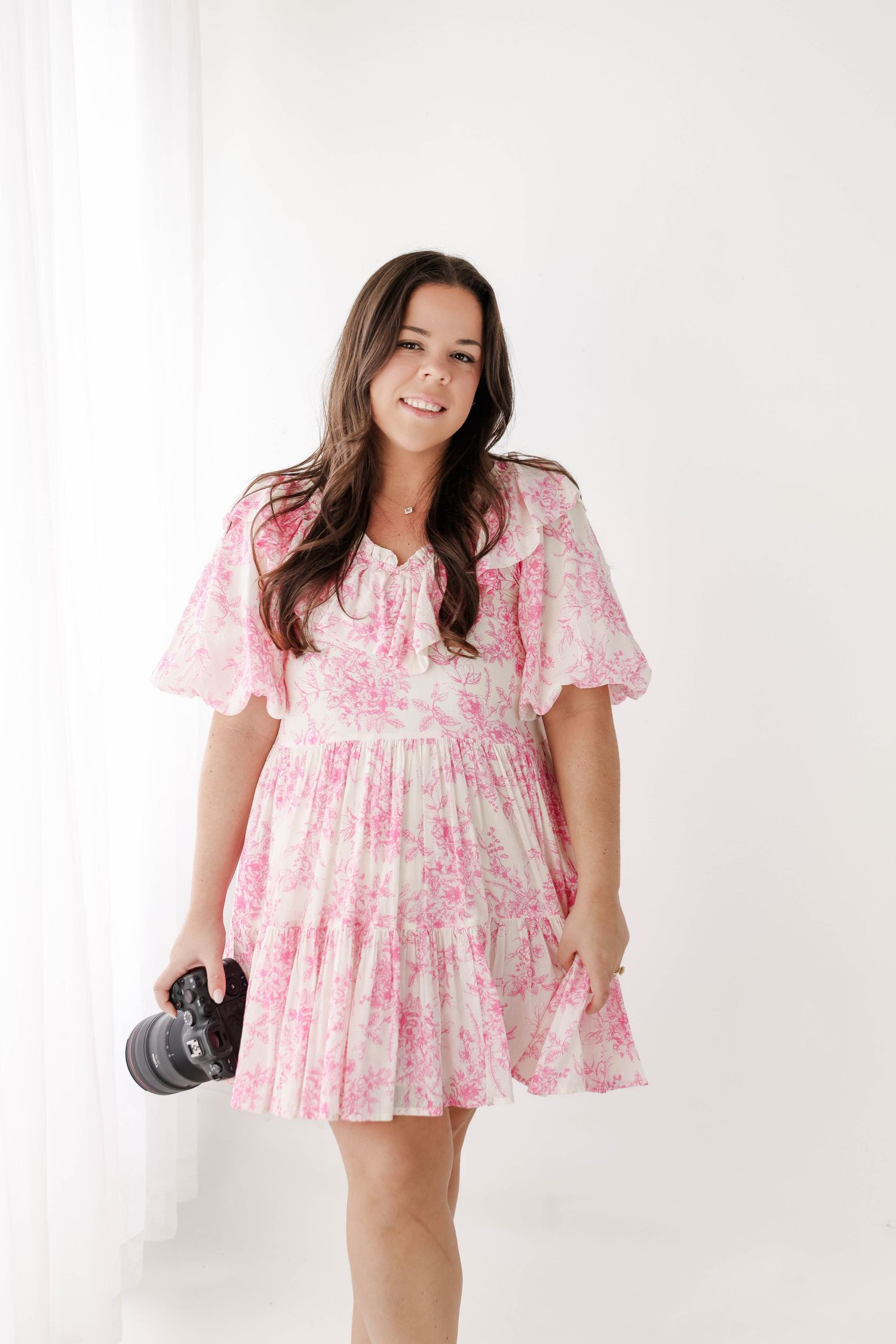 A photographer poses in a feminine pink floral dress with a camera in hand.