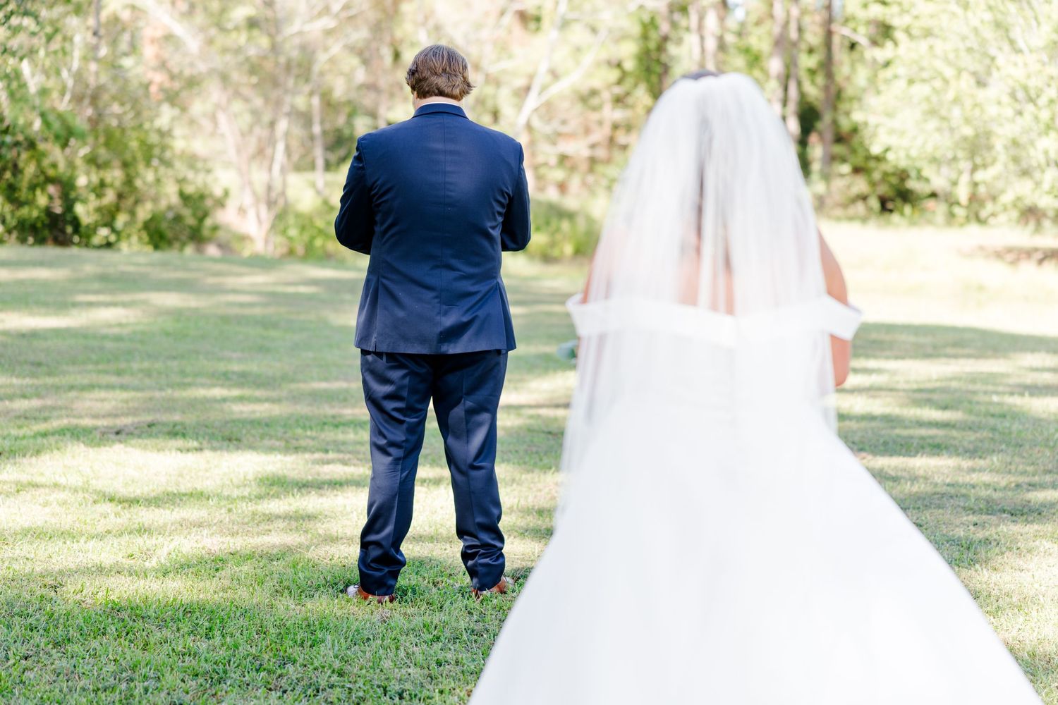 Outdoor wedding scene with bride in white dress and figures in navy suits on grass.