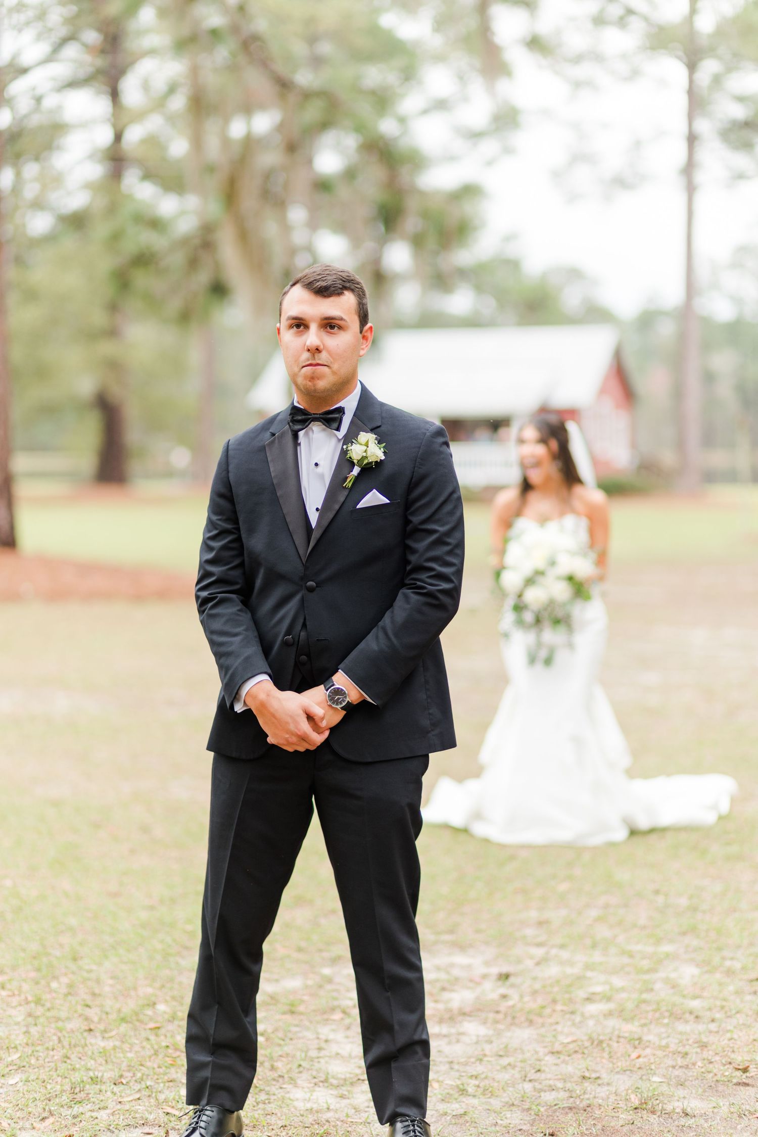 A groom in a classic black tuxedo poses playfully in an outdoor setting with pine trees.