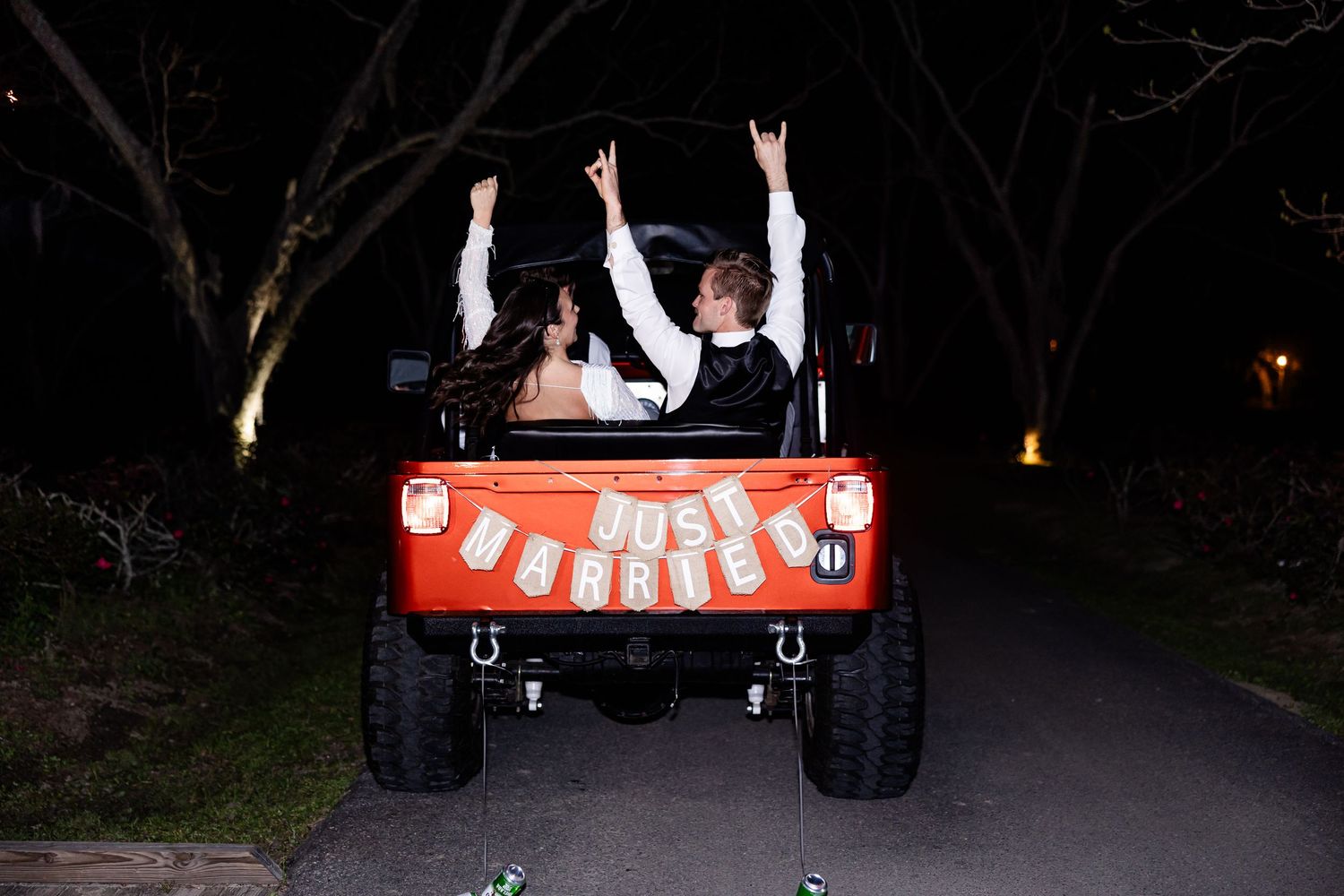 Wedding couple rides in decorated golf cart through dark wooded path at night.
