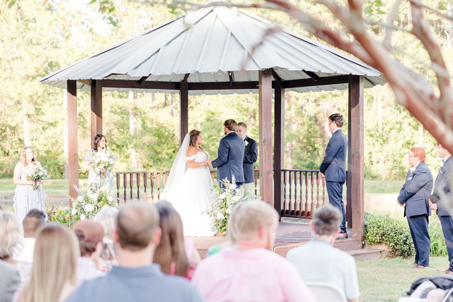 Outdoor wedding ceremony under a wooden gazebo with guests seated on white chairs in a natural setting.