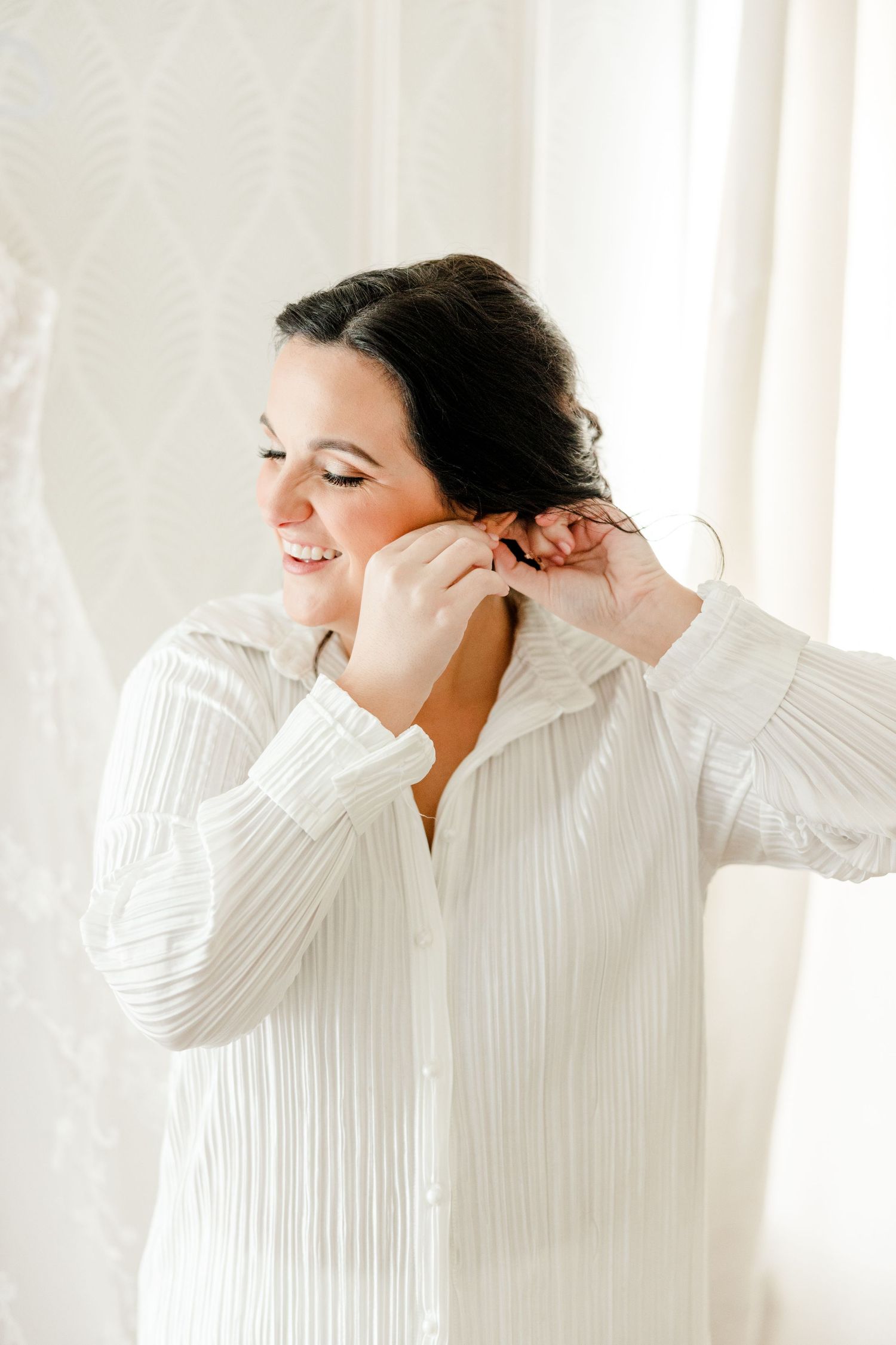 Woman wearing white pleated top smiles while adjusting her hair in a bright studio setting.