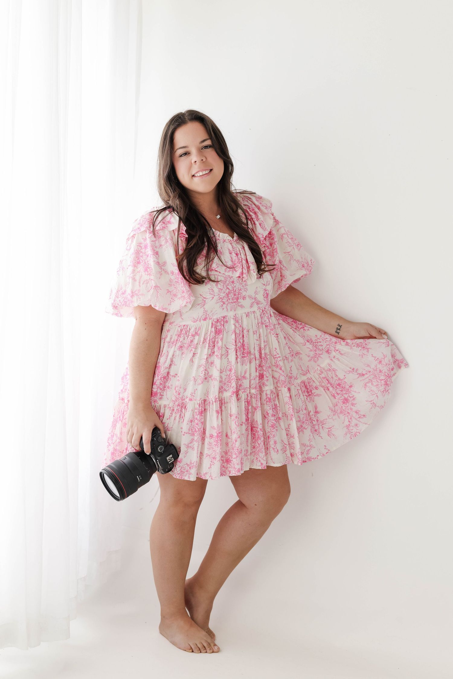 A photographer shows off a flowy pink floral dress while holding a professional camera.
