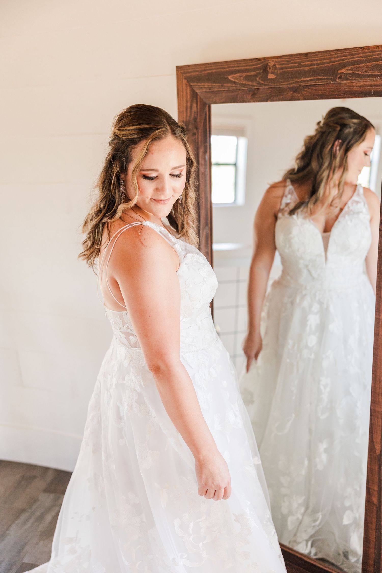 A bride in a lace wedding gown looks down while standing next to a wooden framed mirror.