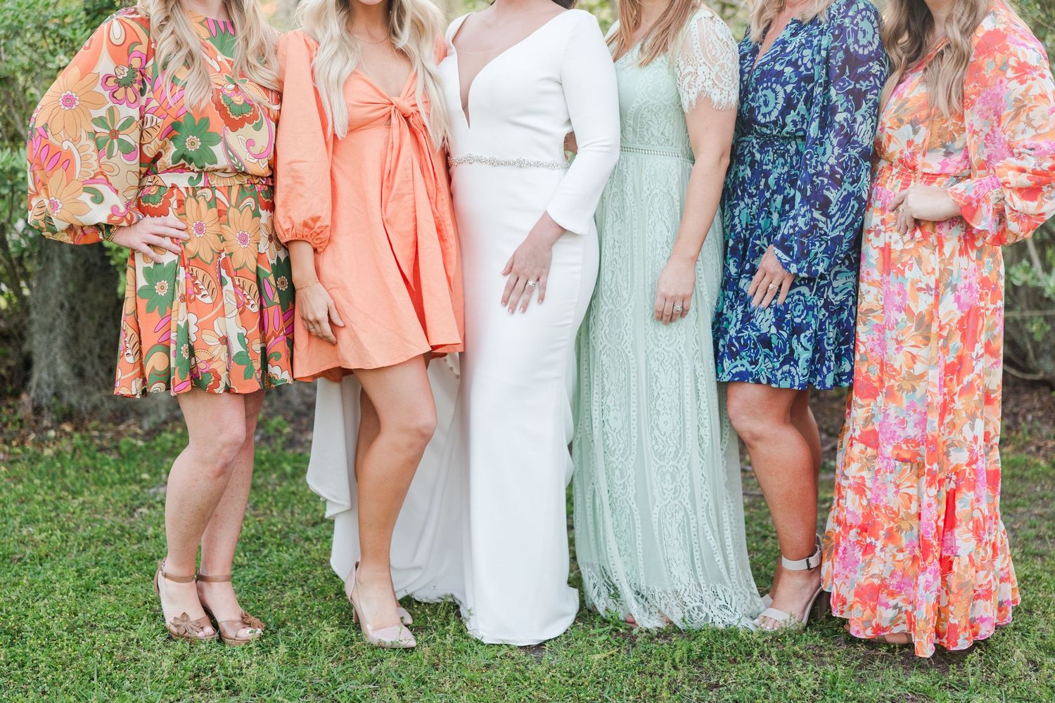 Wedding party members in colorful floral dresses pose together on grass.