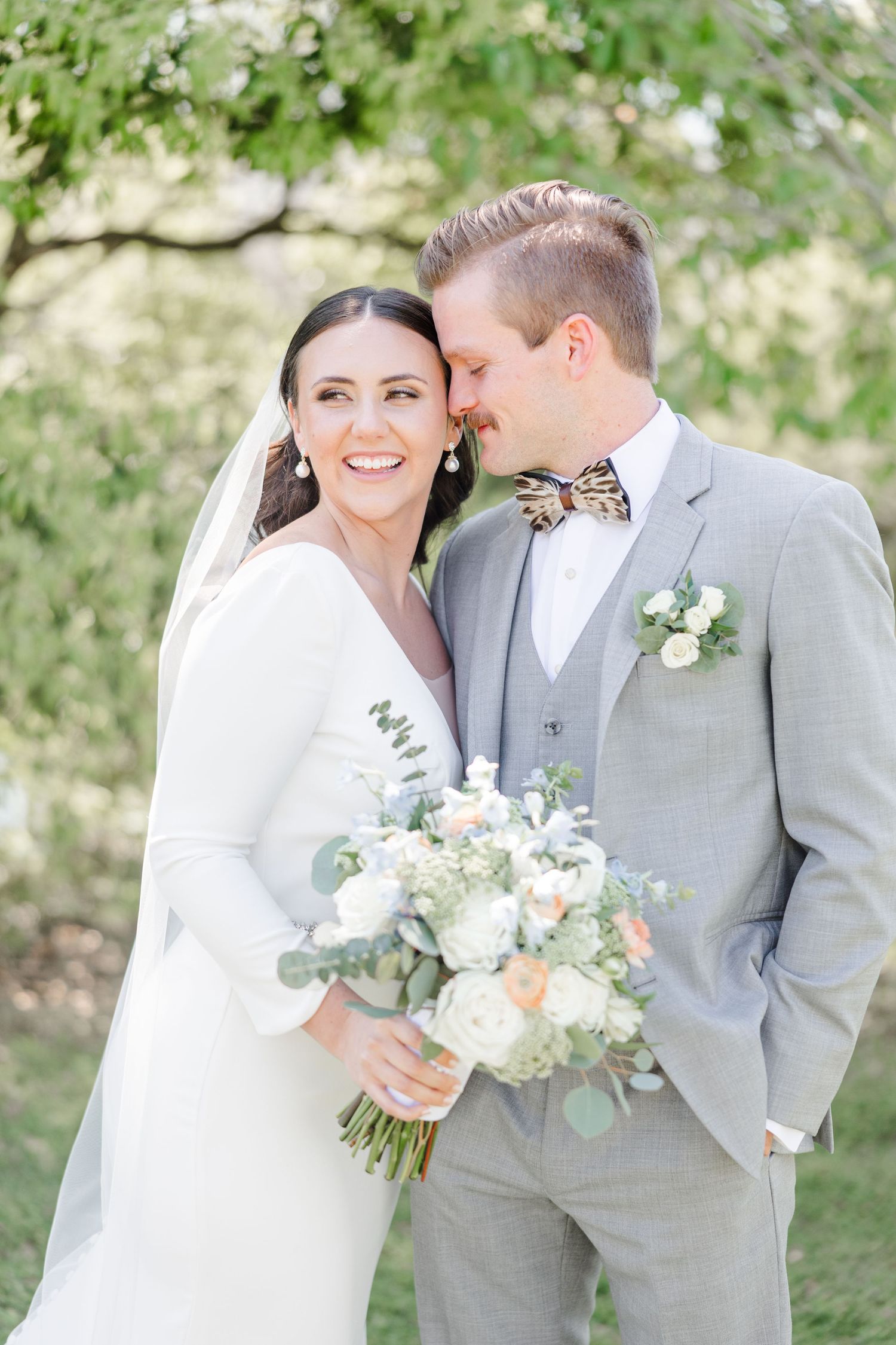 Newlyweds share intimate moment outdoors with white floral bouquet against green foliage.