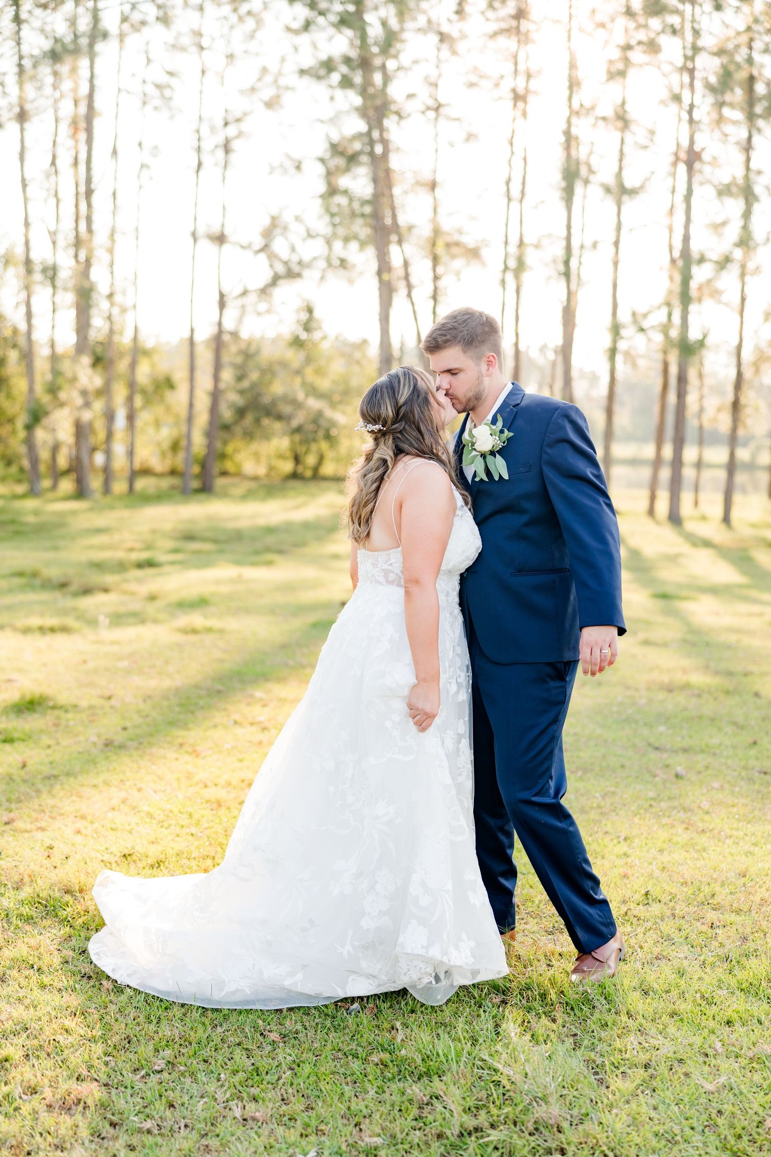 A romantic wedding moment captured in a sunlit forest clearing with pine trees.