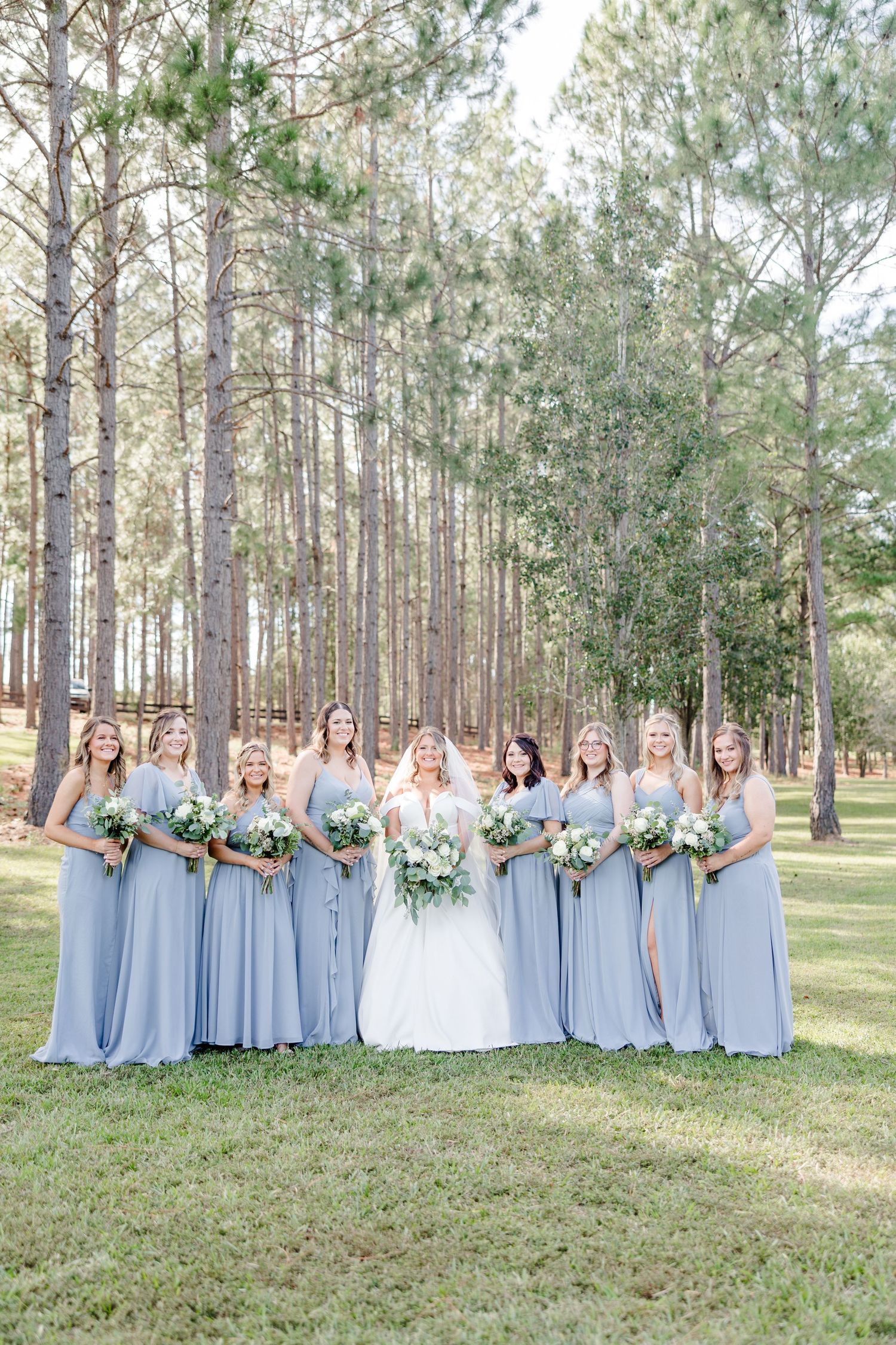 Bridesmaids in light blue dresses pose together in a pine forest setting for wedding photos.