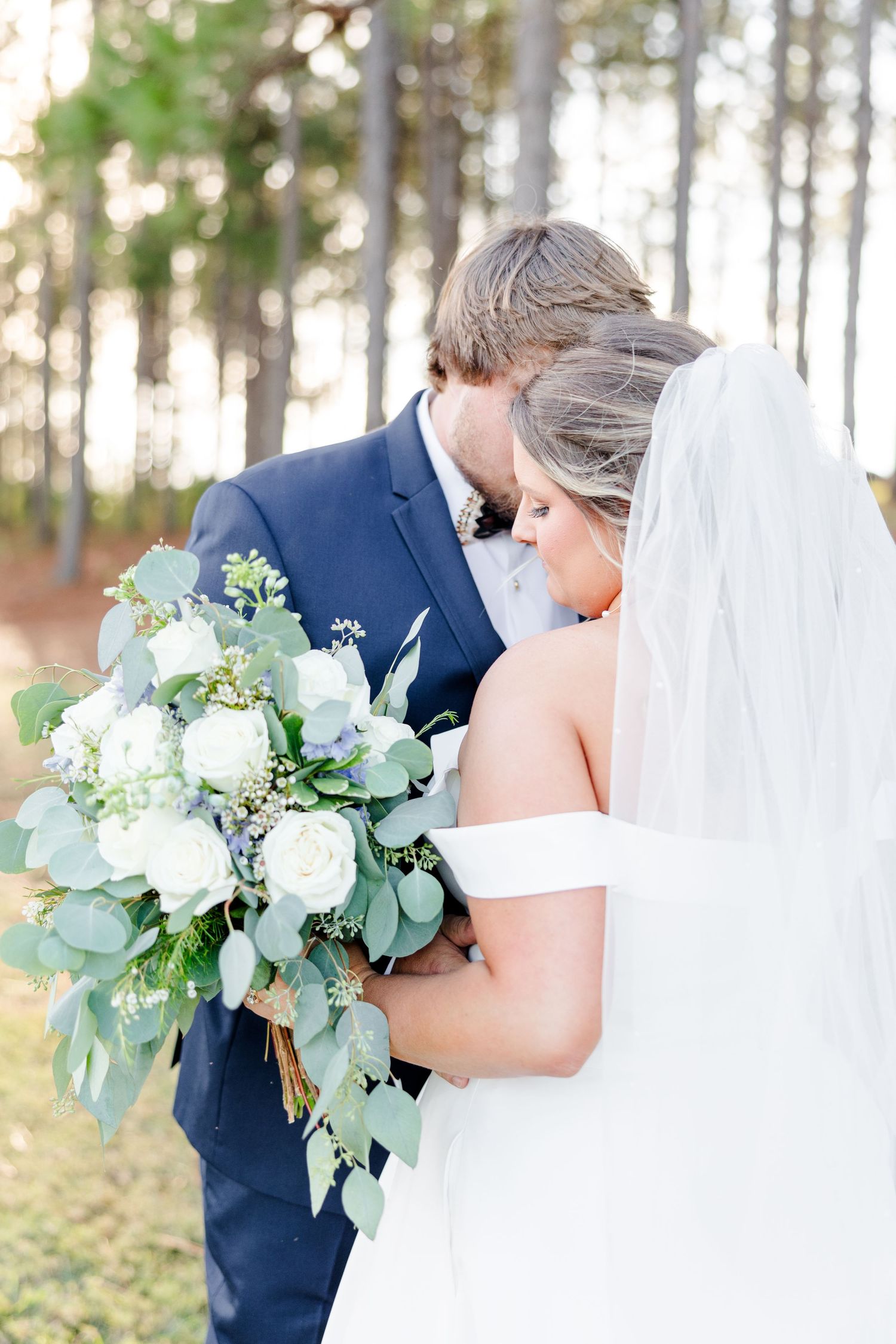 Romantic wedding couple embraces in a sunlit pine forest with white and green floral bouquet.