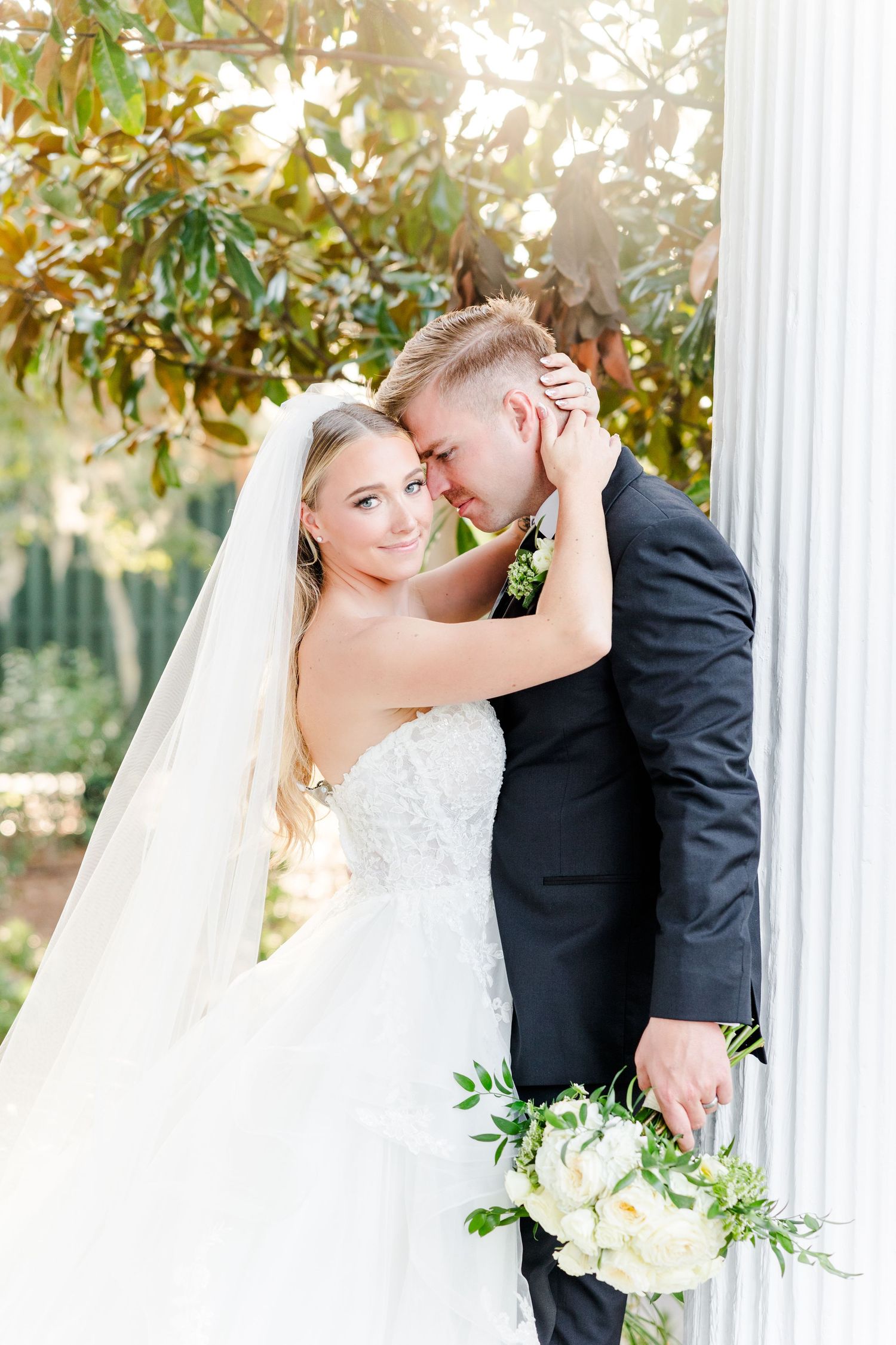 Romantic wedding couple embracing between white columns in an outdoor garden setting.