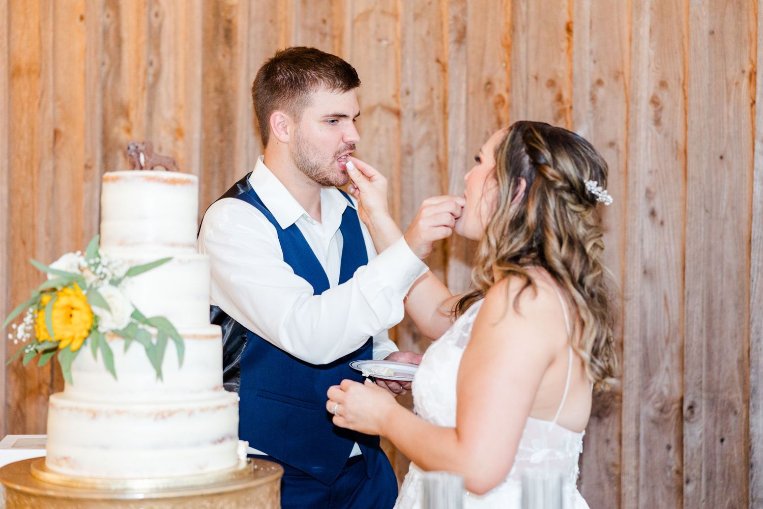 Couple laughs while feeding each other wedding cake in front of wooden wall.