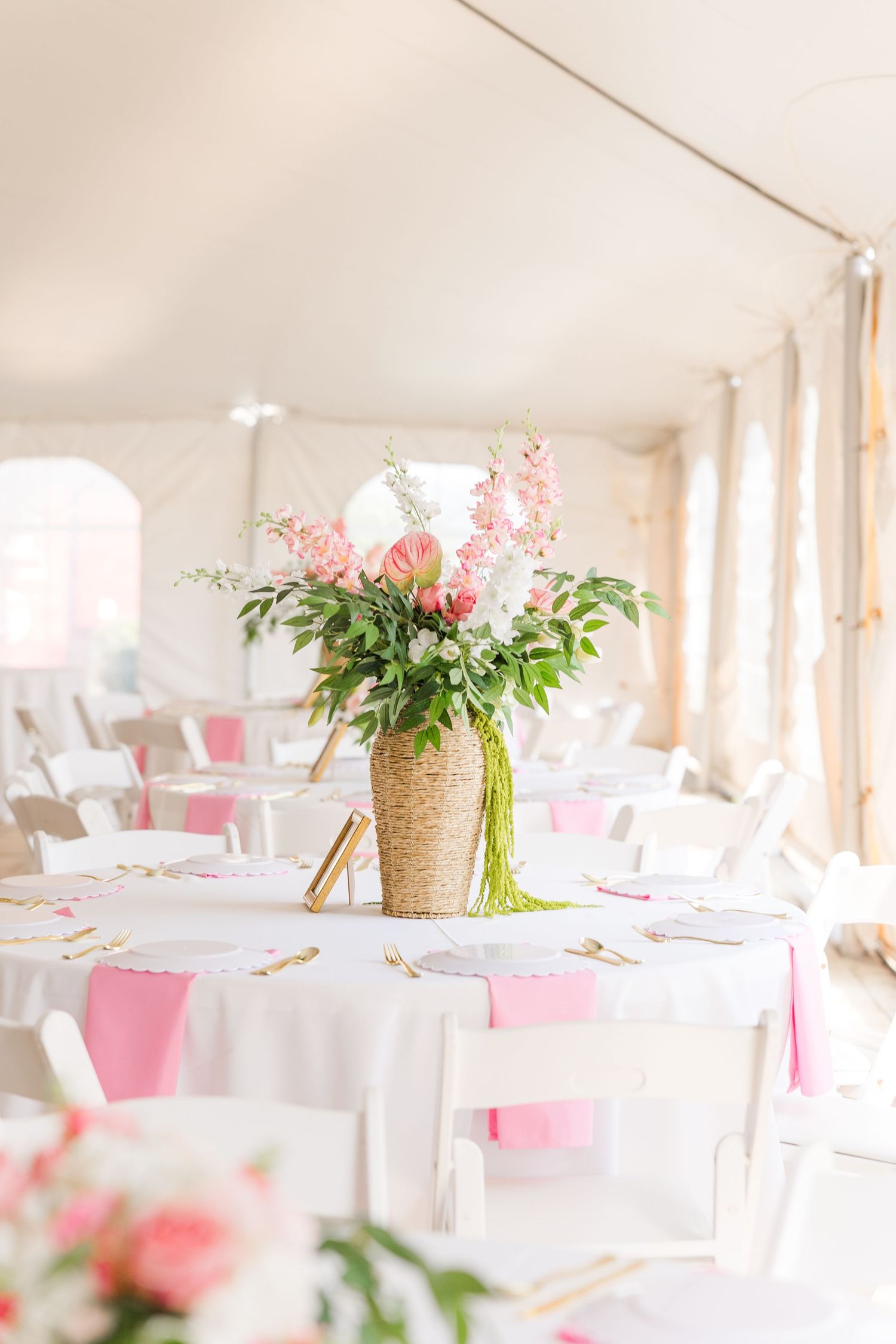 Elegant wedding reception tent with white tables, pink floral centerpieces in wicker vases, and pink ribbon accents.