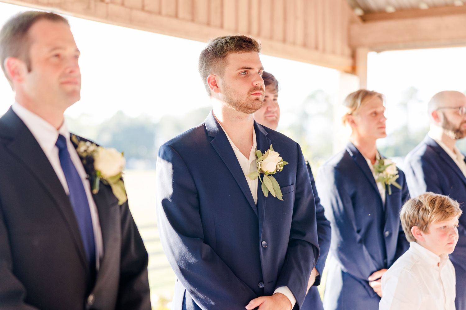 Groomsmen in navy suits stand together during outdoor wedding ceremony.