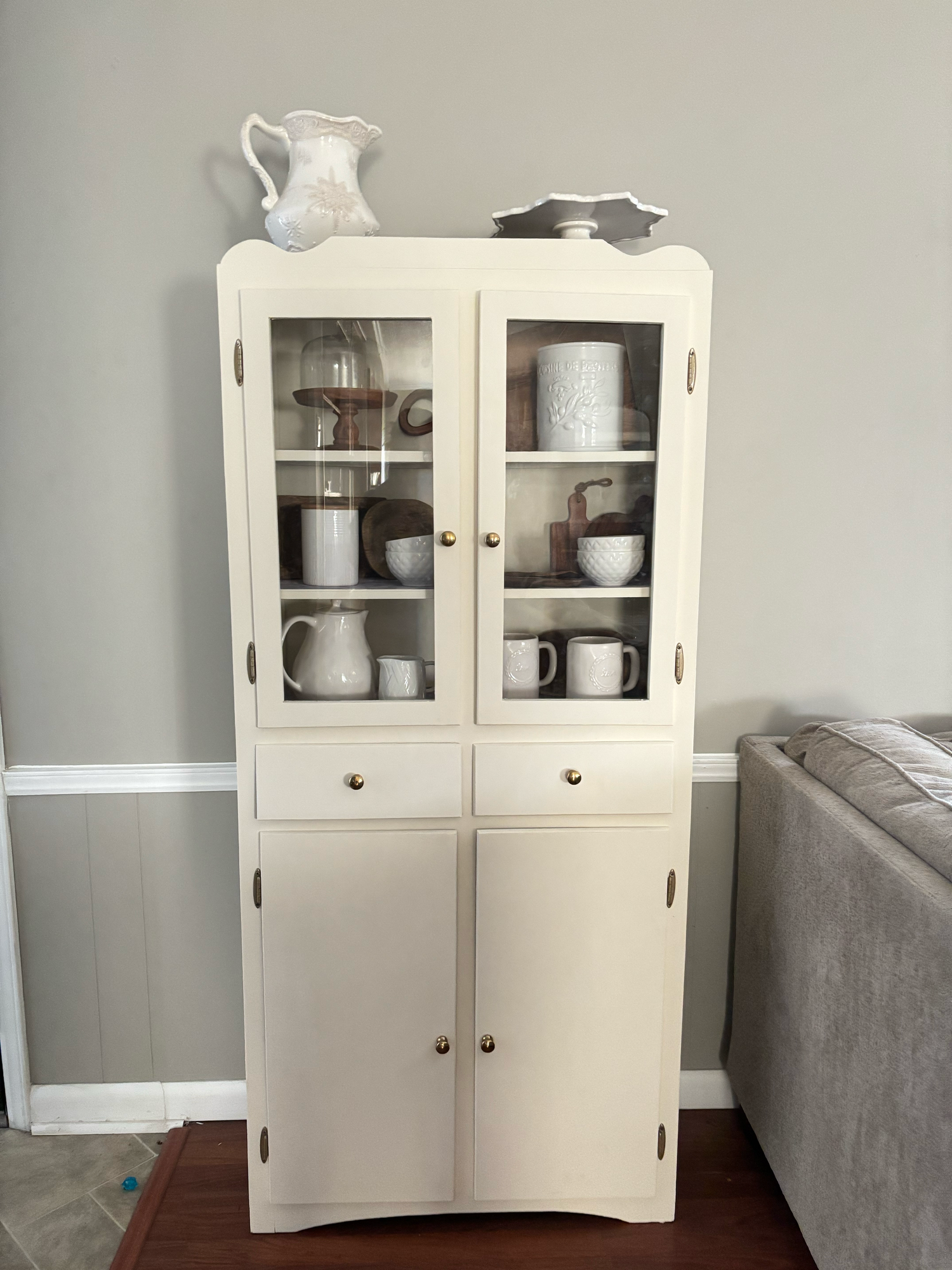 White vintage china cabinet with glass doors displaying ceramic mugs, pitchers, and dishware on wooden shelves.