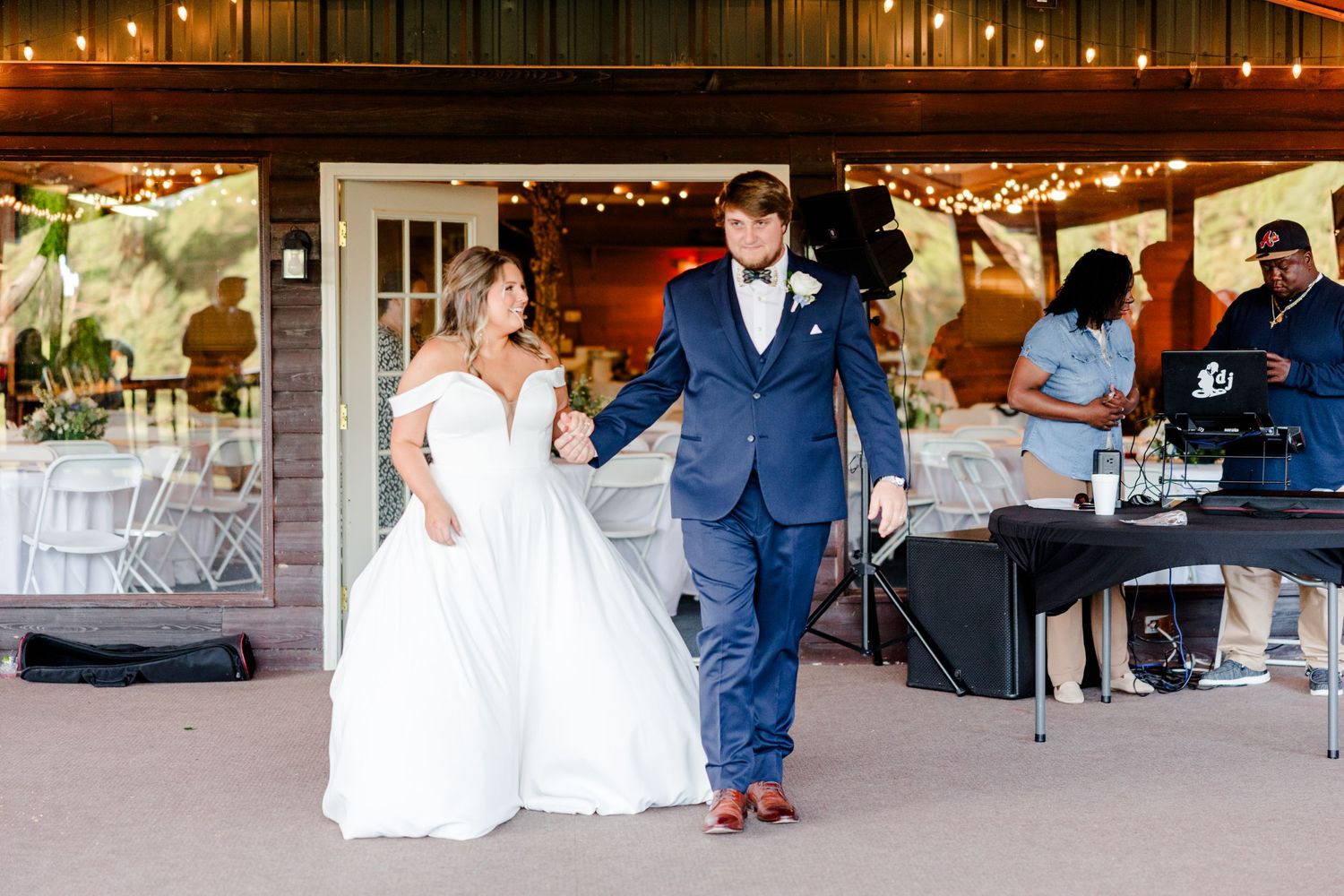 A couple dances together at a wedding reception under warm string lights and wooden beams.