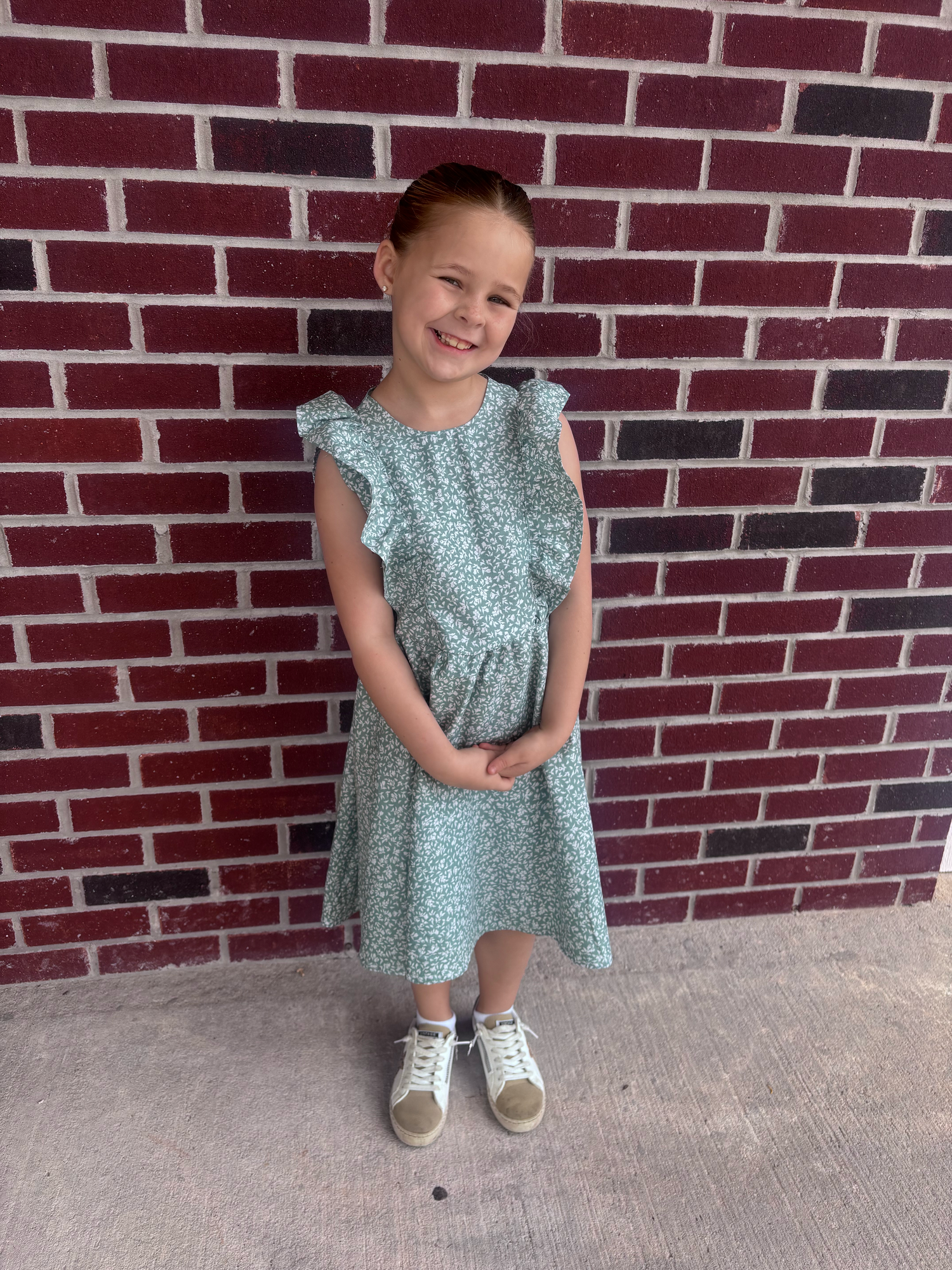 Young girl in a mint floral dress and white sneakers smiling in front of a red brick wall.