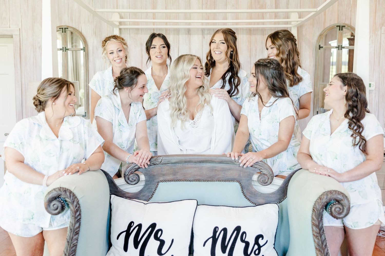 Wedding party wearing matching white robes poses with decorative signs in bright indoor setting.