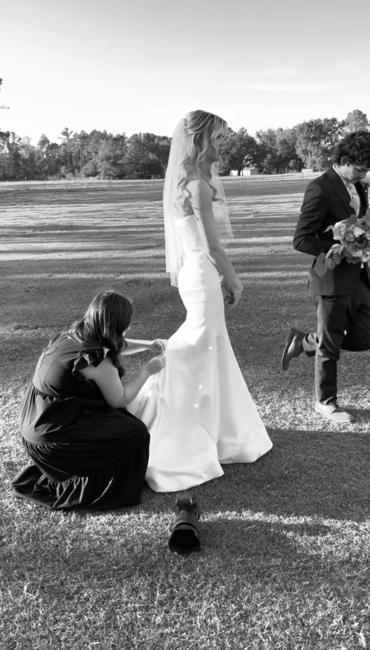 Black and white photo of a bride in white gown being assisted by bridesmaid while groom holds bouquet outdoors.