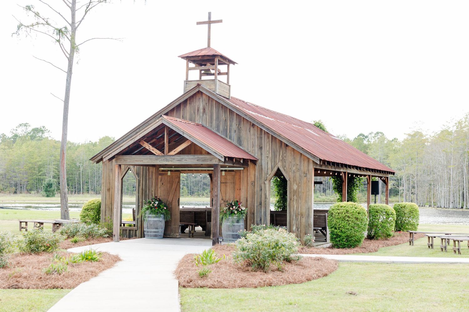 Rustic wooden chapel with red metal roof and cross, surrounded by lush greenery and a paved walkway.