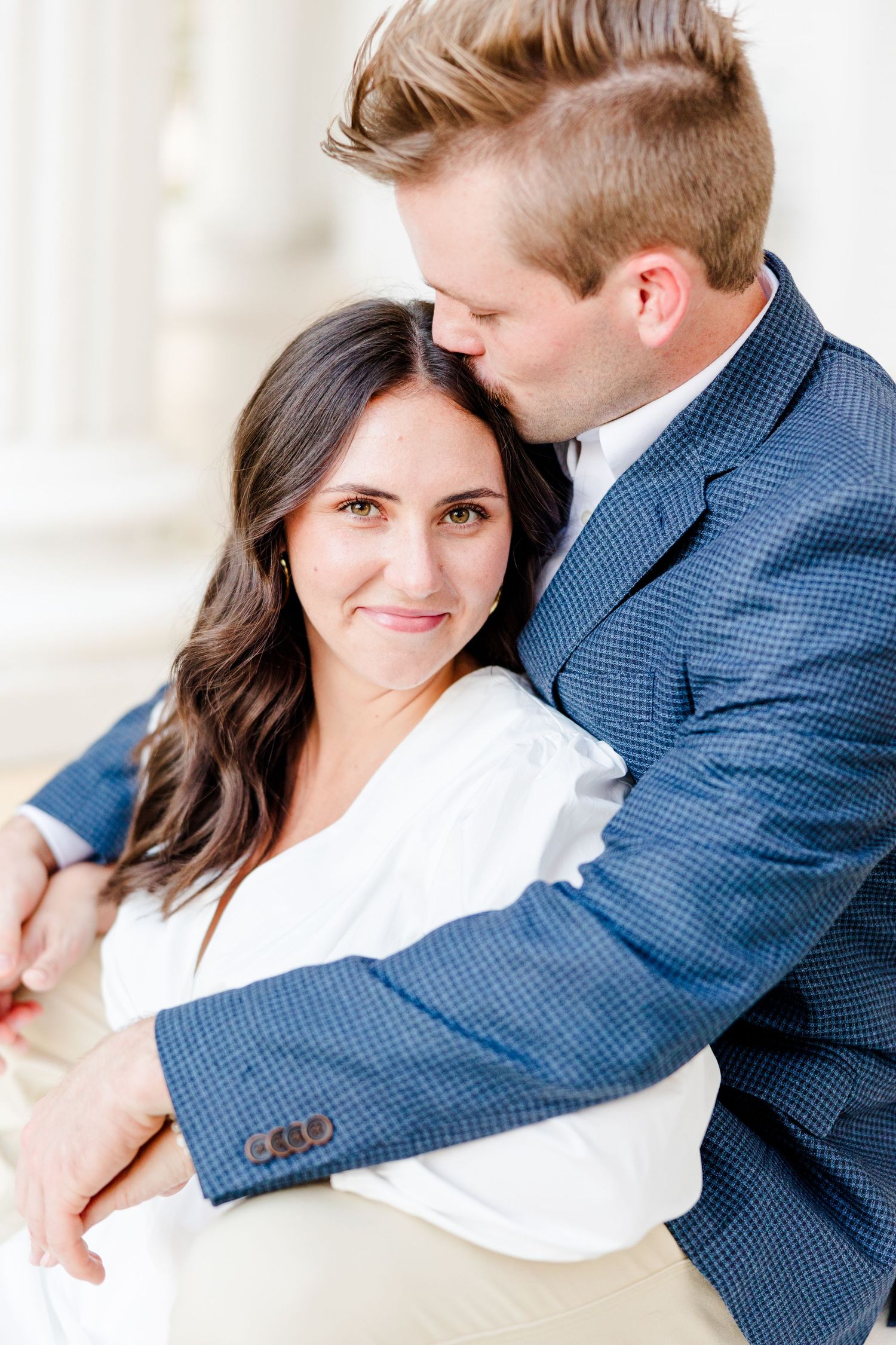 A man in a blue blazer tenderly kisses a dark-haired woman's head while embracing her from behind outdoors.