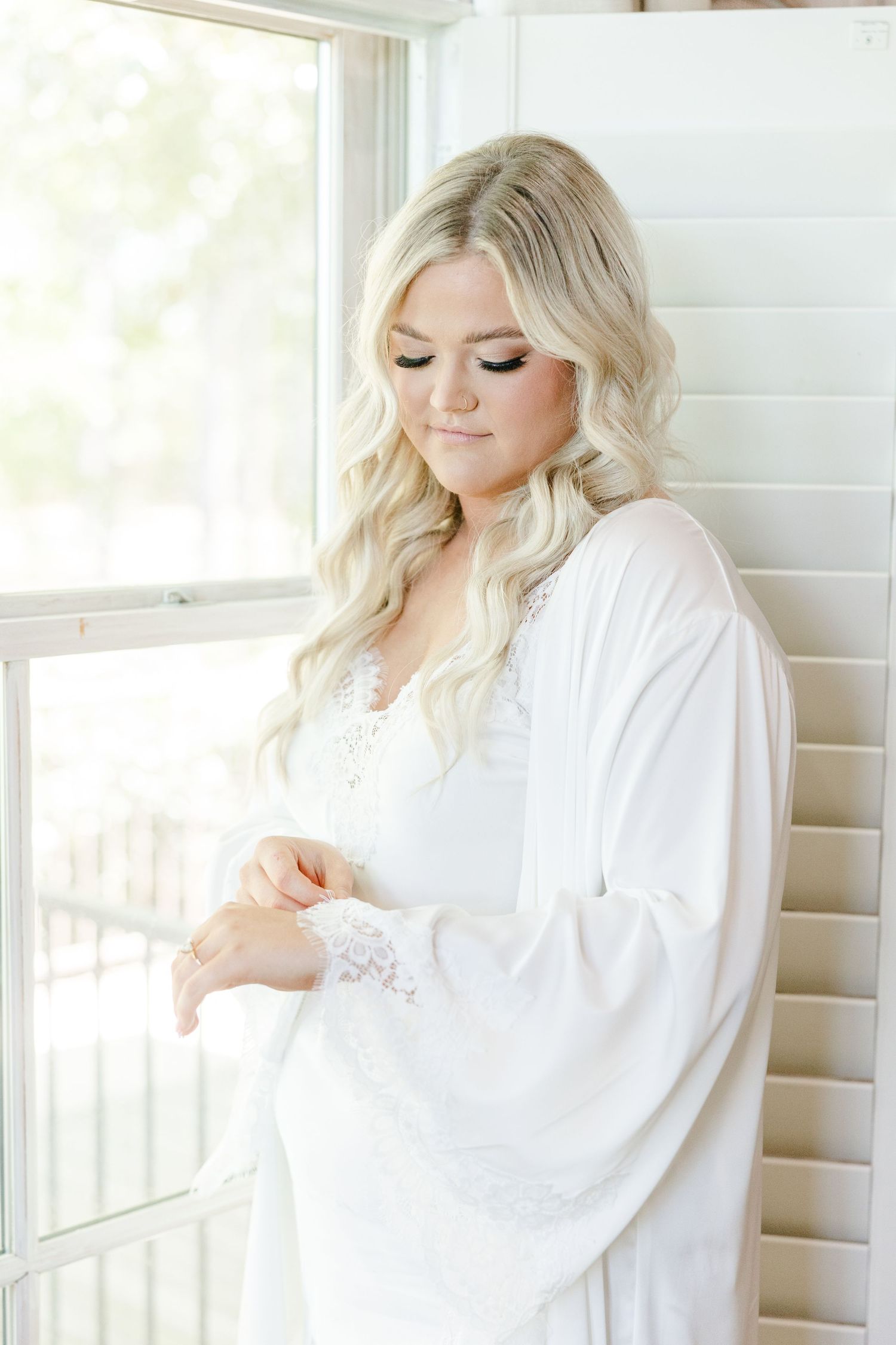 Bride in white silk robe poses gracefully by window during wedding day preparation.