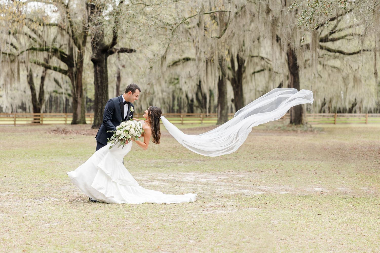 Long flowing bridal veil catches wind dramatically in outdoor wedding portraits.