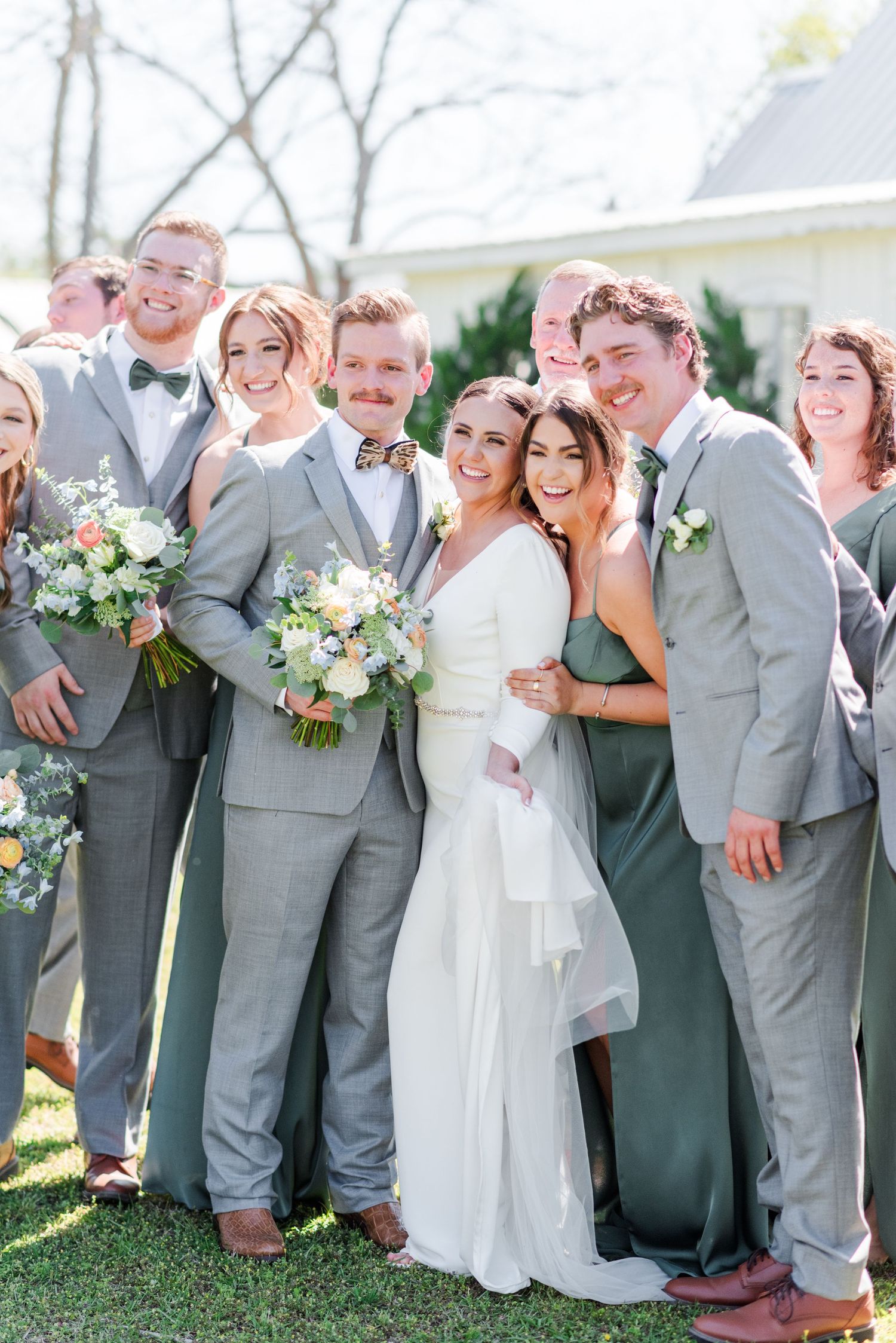 Wedding party in grey suits and sage green dresses laughing together outdoors.