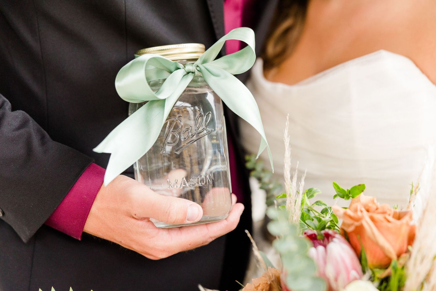 Bride and groom holding a mason jar with green ribbon bow at wedding ceremony.