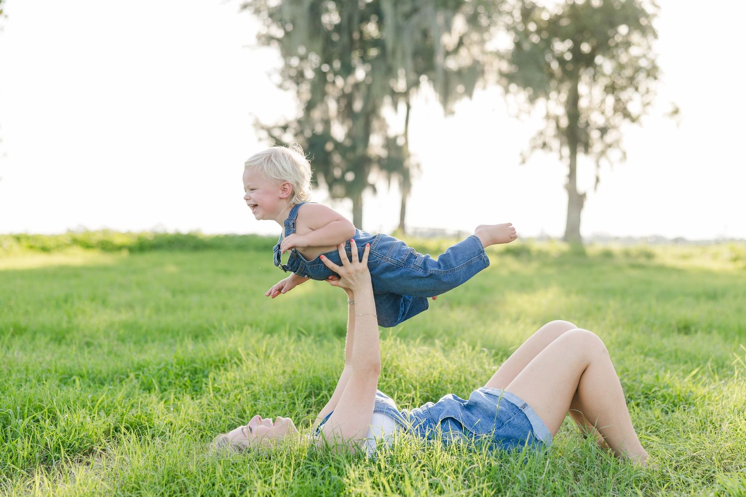 A mother lying on green grass lifts her laughing toddler in the air outdoors near tall trees.