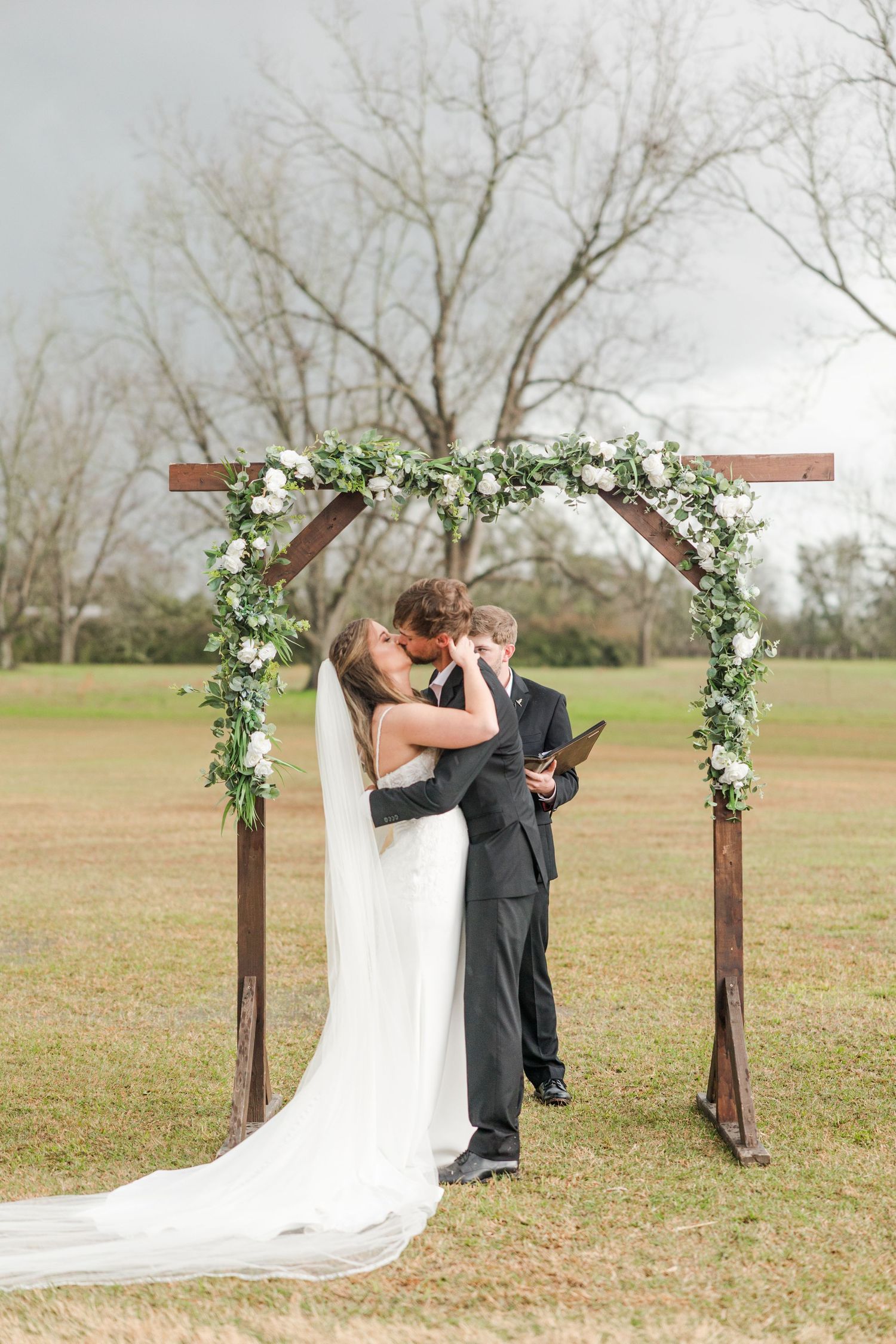 Bride and groom share first kiss under floral arch with white roses and greenery at outdoor wedding ceremony.