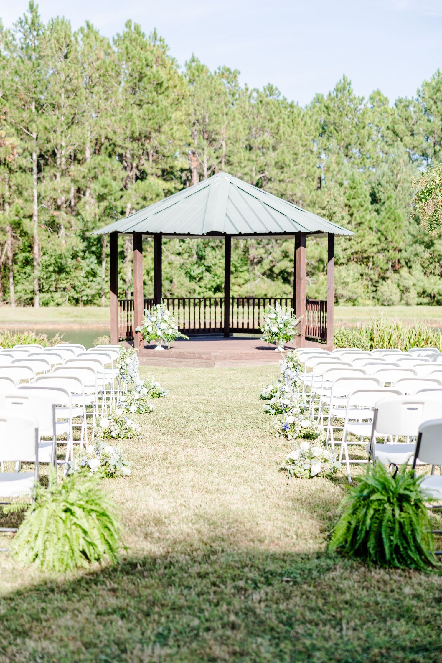 Outdoor wedding ceremony setup with white chairs and gazebo surrounded by towering pine trees.