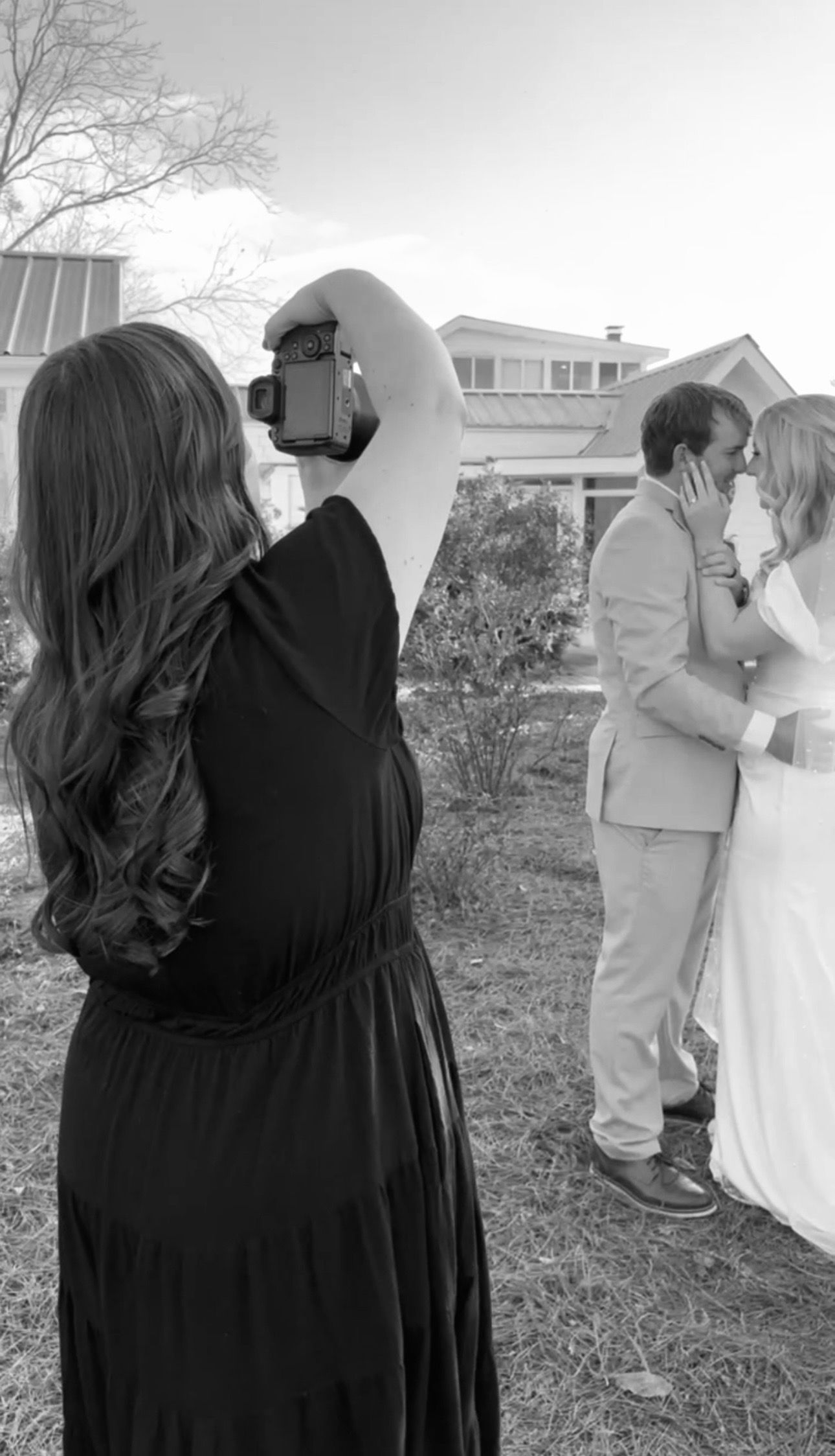 Black and white photo of a wedding photographer capturing a couple kissing outdoors near a house.