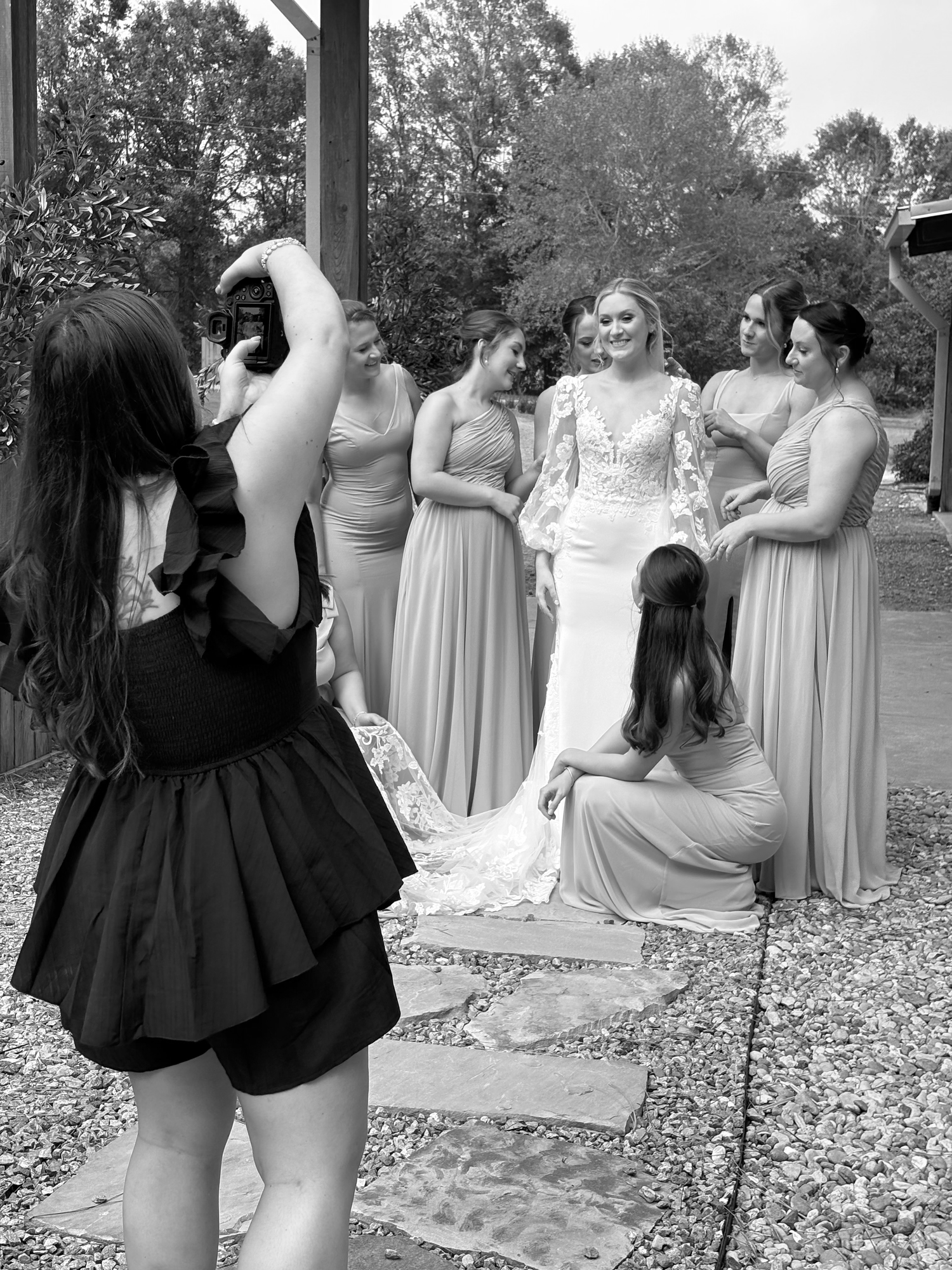 Black and white photo of a bride in a lace gown posing with bridesmaids while a photographer captures the moment.