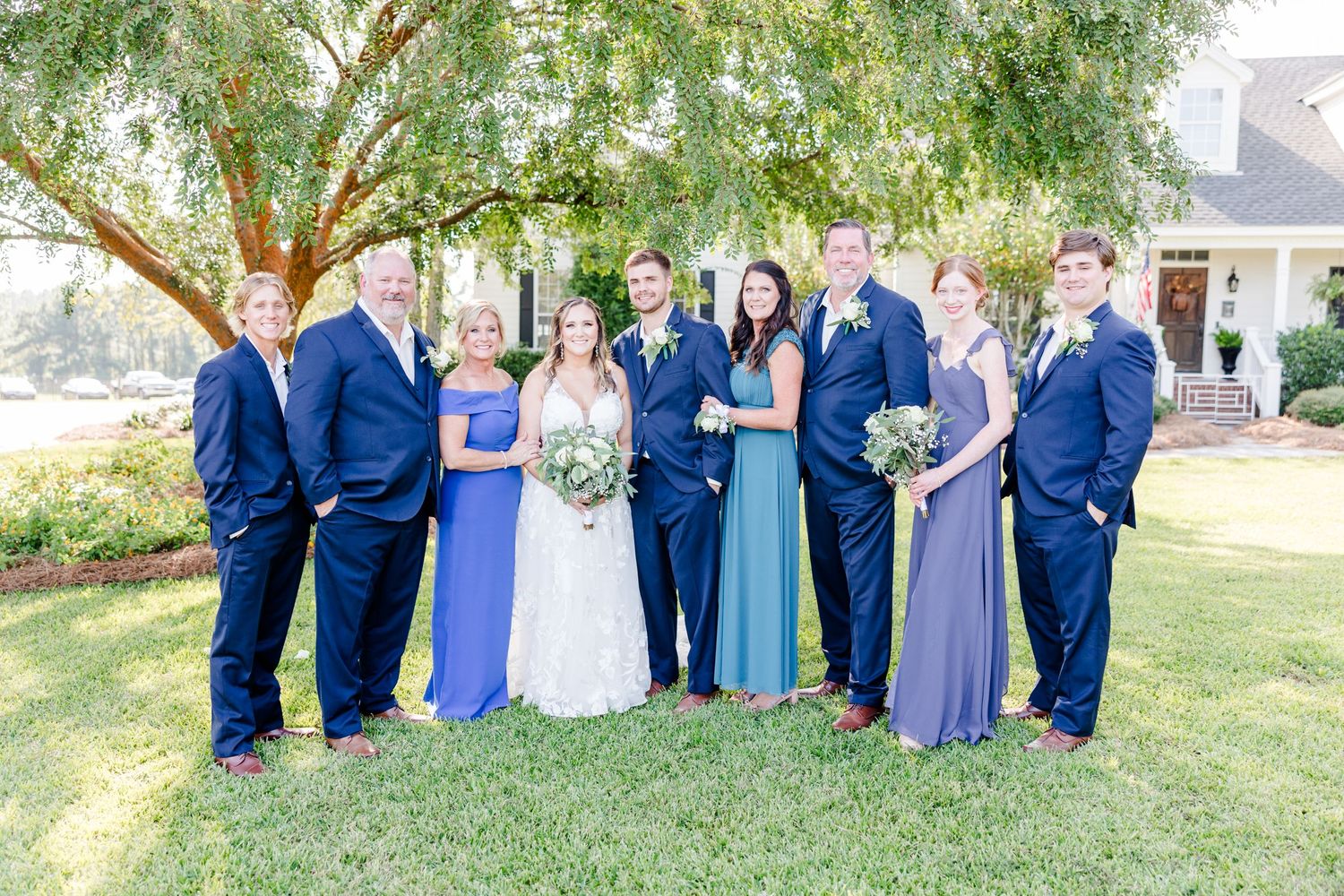 Wedding party gathers under trees for outdoor group photographs.