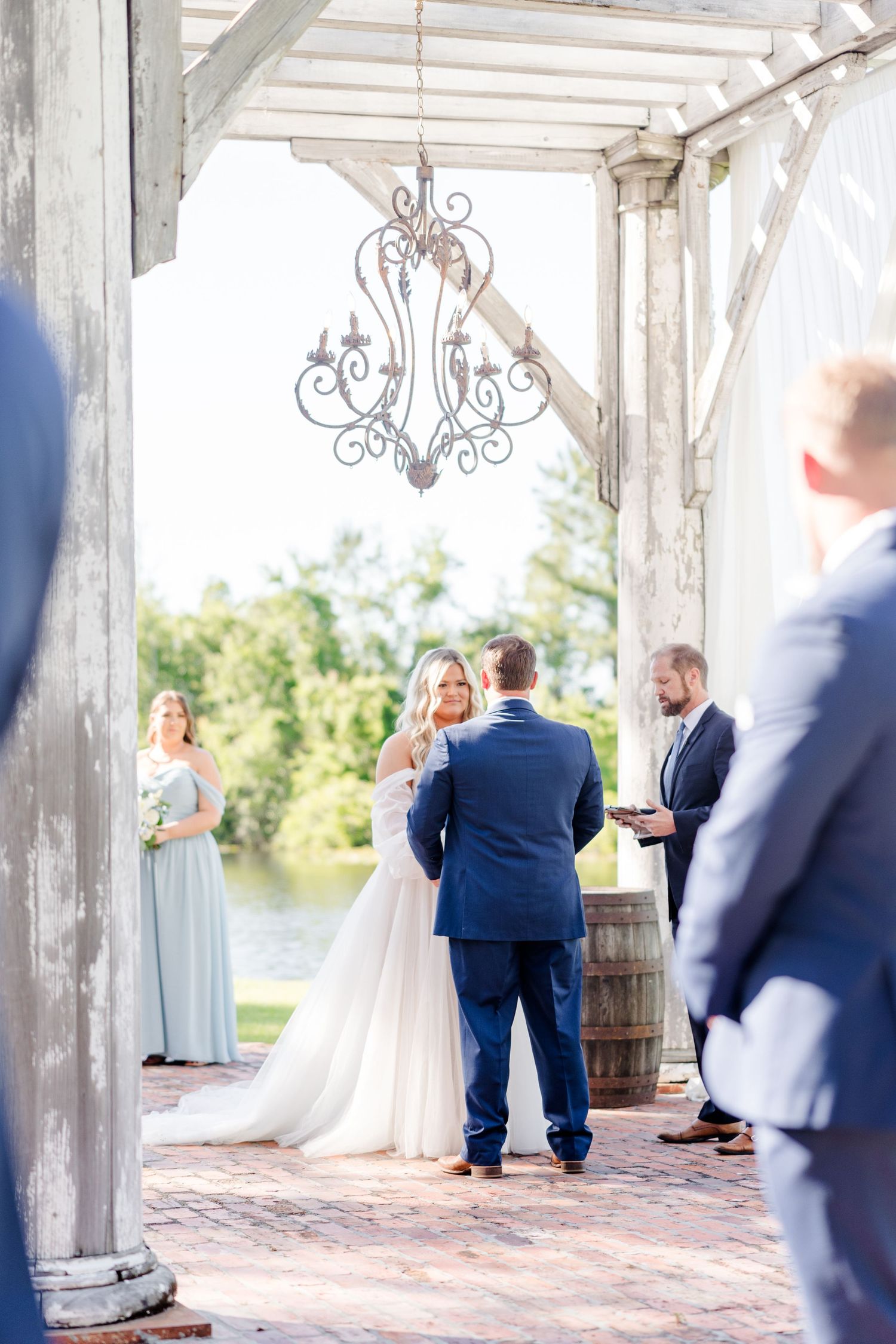 Outdoor wedding ceremony under a white pergola with ornate chandelier and waterfront view.